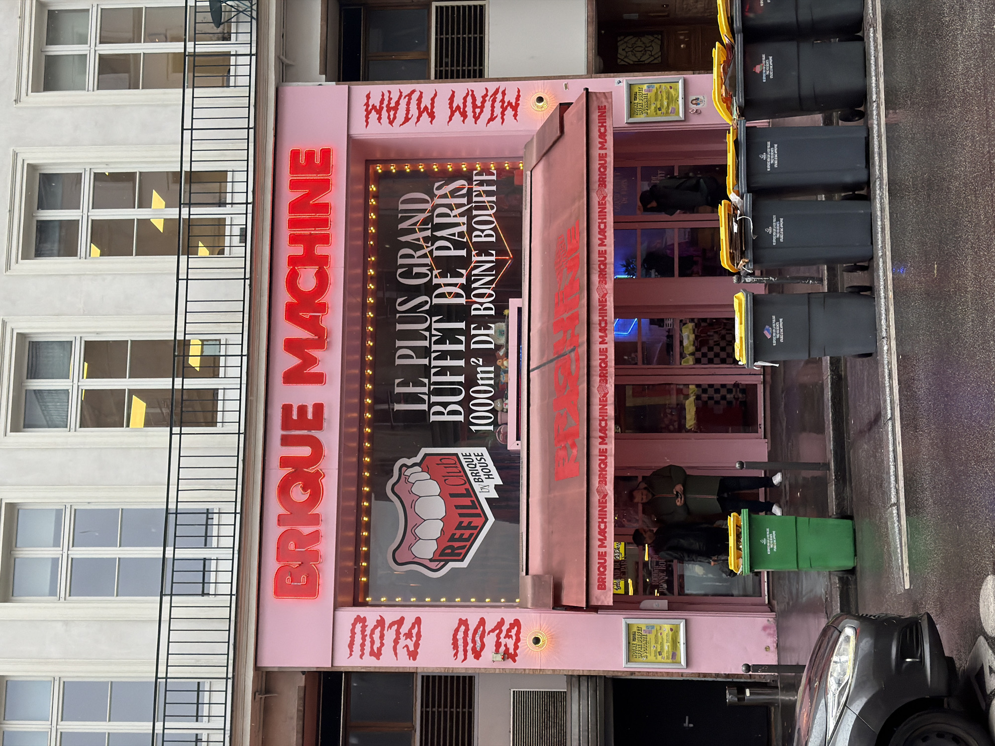 The image depicts the facade of "Brique Machine", a Parisian restaurant. The building is light pink and has windows on the left side. The restaurant's sign reads "BRIQUE MACHINE" in large red letters above an advertisement. The advertisement is for "LE PLUS GRAND BUFFET DE PARIS 1000m² DE BONNE BOUFFE" and includes an image of teeth. The restaurant entrance is located to the right, visible through the window, and has a black-and-white checkered pattern. Several people are standing in front of the restaurant, including two men and a woman looking at the phones. Several black trash containers are located to the right of the restaurant entrance. A green trash container is also located to the right, along with a grey car. The text "MIAM MIAM" is written in red above the restaurant's sign. The restaurant is located in Paris, France.
