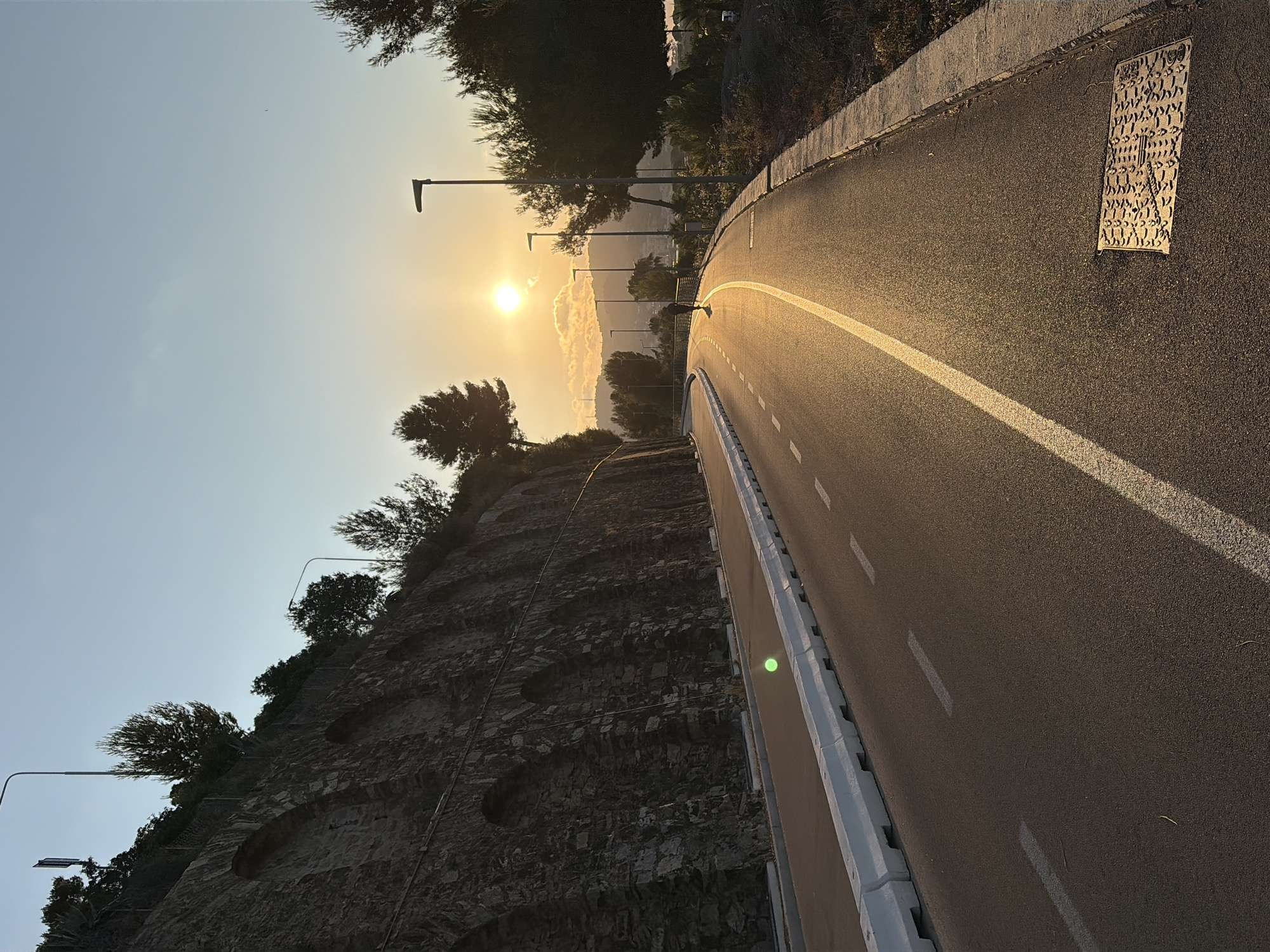 The image shows a road with a bike path on the left, bordered by a stone wall and some vegetation. The road is illuminated by the golden light of a sunset. A lone figure is seen cycling on the road. In the background, there are trees and hills, and the sky is clear blue with some clouds. The scene is peaceful and serene. The location is Imperia, Italy, along the scenic coastal road, likely the Lungomare degli Aranci or a similar coastal path.