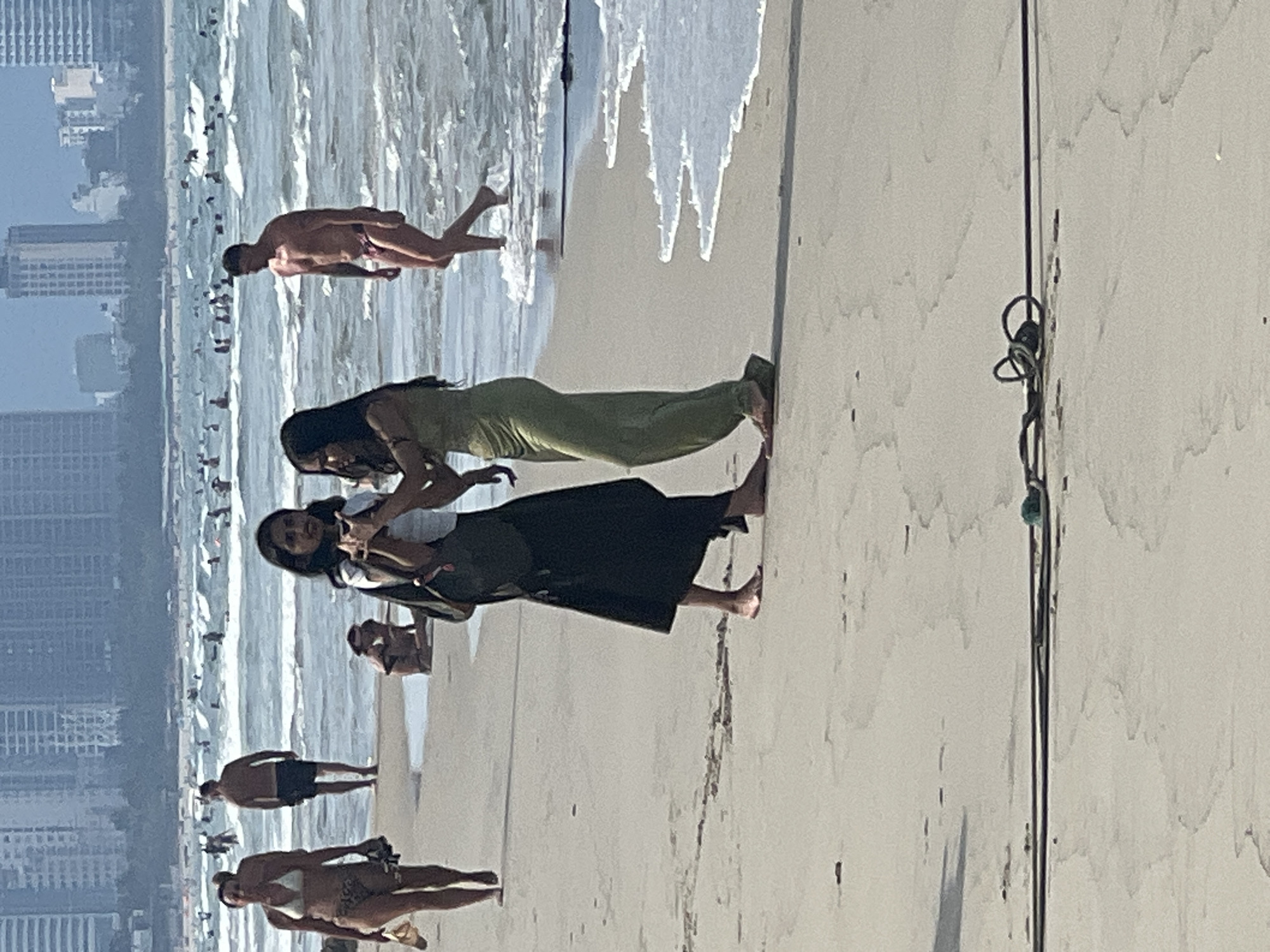 This media file shows a beach scene in Da Nang, Vietnam. The image is oriented vertically, with the ocean and sky appearing on the left and the sandy beach on the right. Several people are visible, some in the water and others on the beach.
In the foreground, three individuals are interacting. Two women, one in a lime green dress and the other in a dark outfit, are positioned close together on the sand. The woman in the green dress appears to be reaching out or touching the other woman. Further in the background, a man in swim trunks is standing on the sand, facing the ocean. In the far background, more people are scattered along the shoreline and in the water, with buildings visible on the horizon.
The sun appears to be high in the sky, suggesting it is likely midday. The weather seems to be clear and sunny, with waves gently breaking on the shore. The sand is light in color, and there are some small dark marks on it. A thin dark cord with a loop is visible on the sand in the right portion of the image. The overall atmosphere seems relaxed and typical of a beach day.
