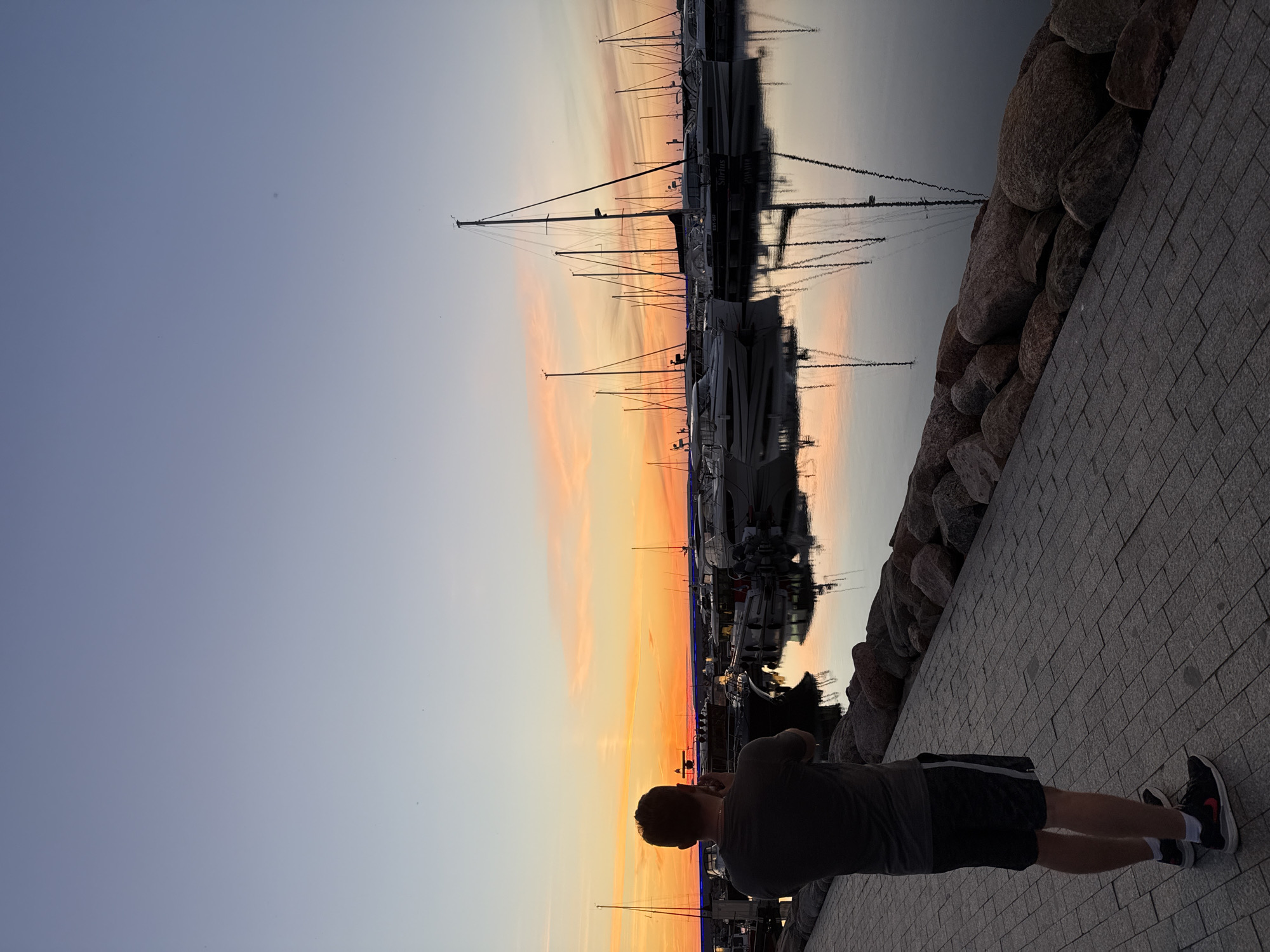The image shows a man standing on a paved walkway at a marina during sunset. He is facing away from the camera, holding a camera up to his face as if taking a photograph. He is wearing a dark t-shirt, dark shorts with a white stripe, and athletic shoes. The marina is filled with numerous sailboats and yachts moored in the water. The sky is a gradient of blue and orange, indicating a sunset. The water reflects the sky and the boats, creating a mirror-like effect. The walkway is made of interlocking gray pavers, and a line of large rocks separates the walkway from the water. The scene appears to be the Tallinn Bay waterfront in Tallinn, Estonia, a common location for sunset views over the Baltic Sea.
