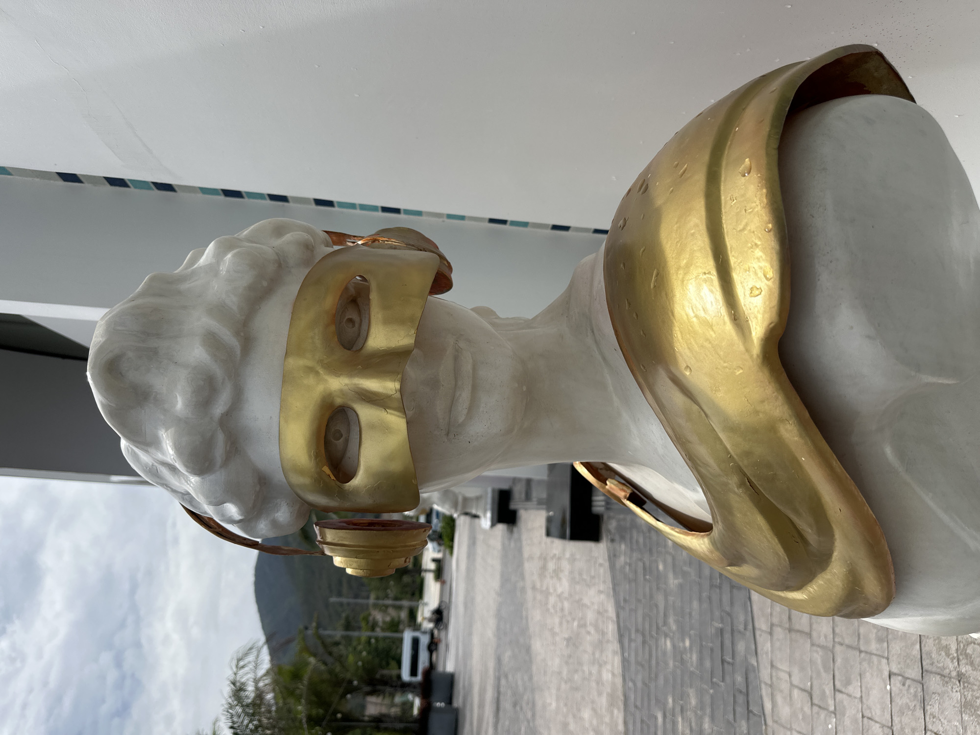A marble bust wearing a golden mask and headpiece is shown. The bust is white and features the head and upper torso of a classical figure with curly hair. A golden mask covers the eyes of the bust, and a golden headpiece is draped over the head and shoulders. The bust is located outdoors, likely on a patio or walkway. In the background, there is a glimpse of a street with cars and buildings. The photo was taken outdoors in Bac Nha Trang Ward, Vietnam.