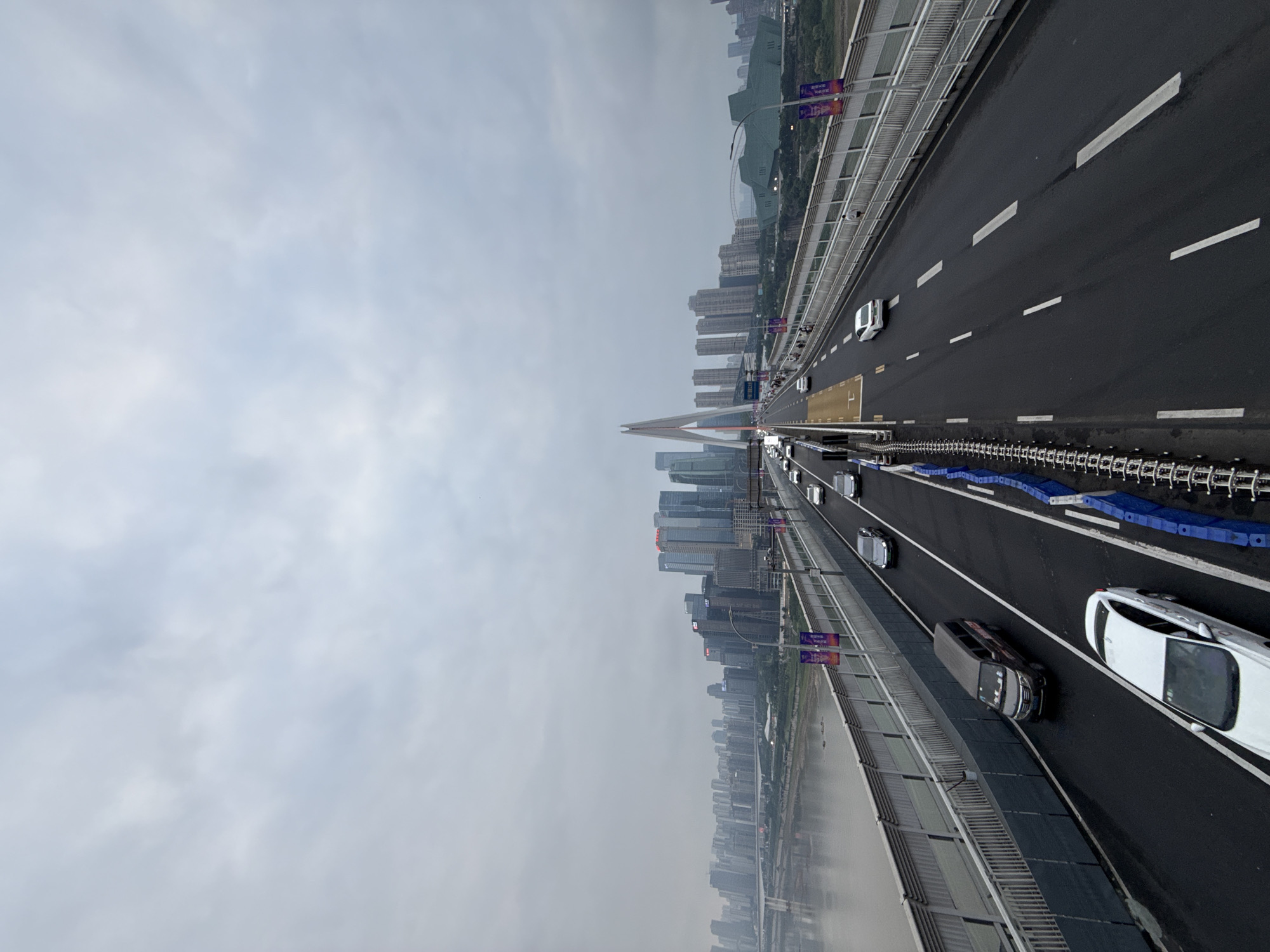 The image depicts a view of the Chongqing Yangtze River Bridge in Yuzhong District, China, on a cloudy day. The bridge spans a wide river, with a city skyline visible in the distance. The bridge is carrying vehicular traffic, with several cars and a van visible on the roadway. Pedestrians are also present on a walkway adjacent to the road. The bridge is a cable-stayed design, with a prominent white pylon rising from the center. The sky is overcast, and the light is diffused, giving the scene a somewhat muted appearance. The water in the river is calm, reflecting the gray sky. The overall impression is one of a busy urban environment, with the bridge serving as a vital link between different parts of the city.