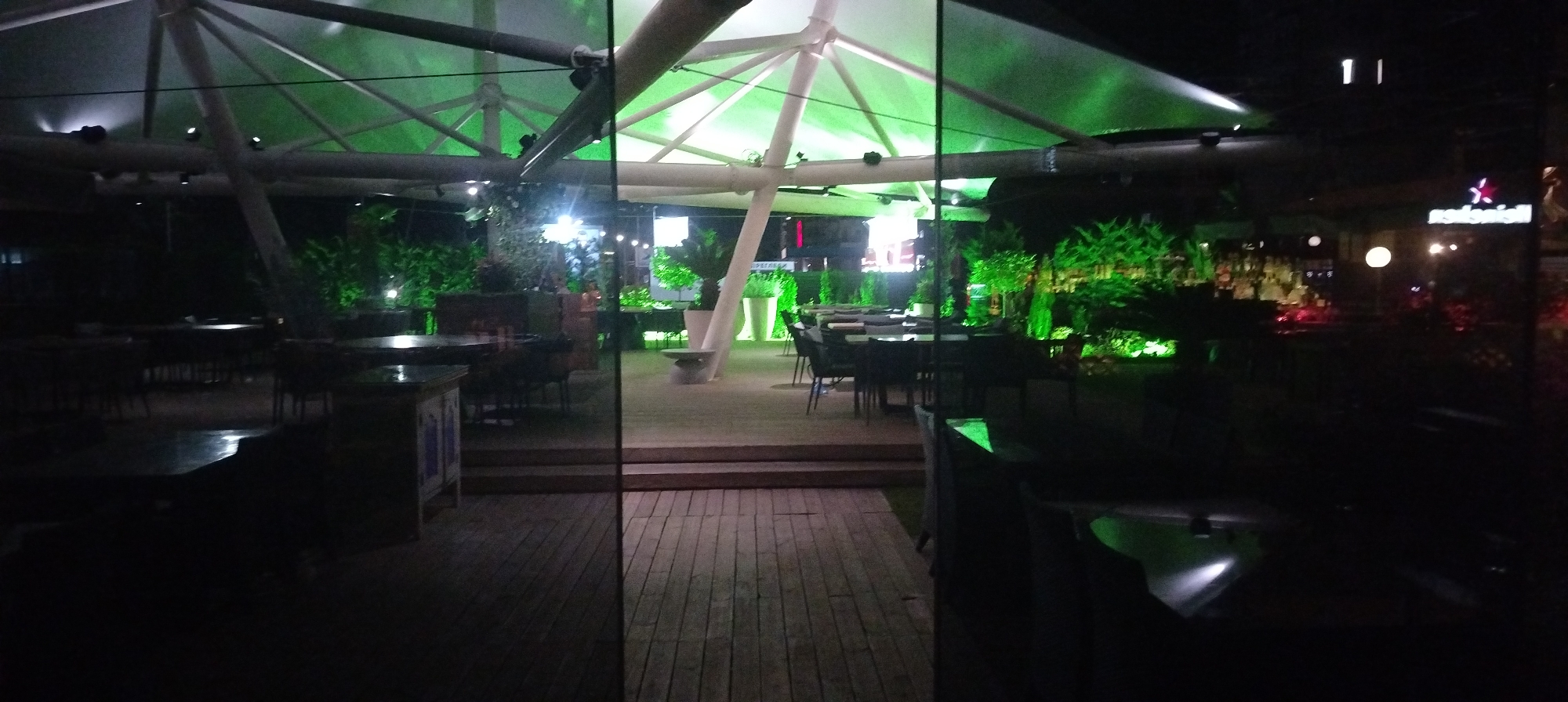 The image depicts the outdoor seating area of a restaurant or bar at night, illuminated by green and white lights. Tables and chairs are set up on a wooden deck and a paved area. In the background, a bar area with bottles and lights is visible. The establishment appears to be in an urban setting, potentially Sofia, Bulgaria, as indicated by the presence of signage which, though partially obscured, includes Cyrillic script consistent with Bulgarian. The scene is devoid of people.