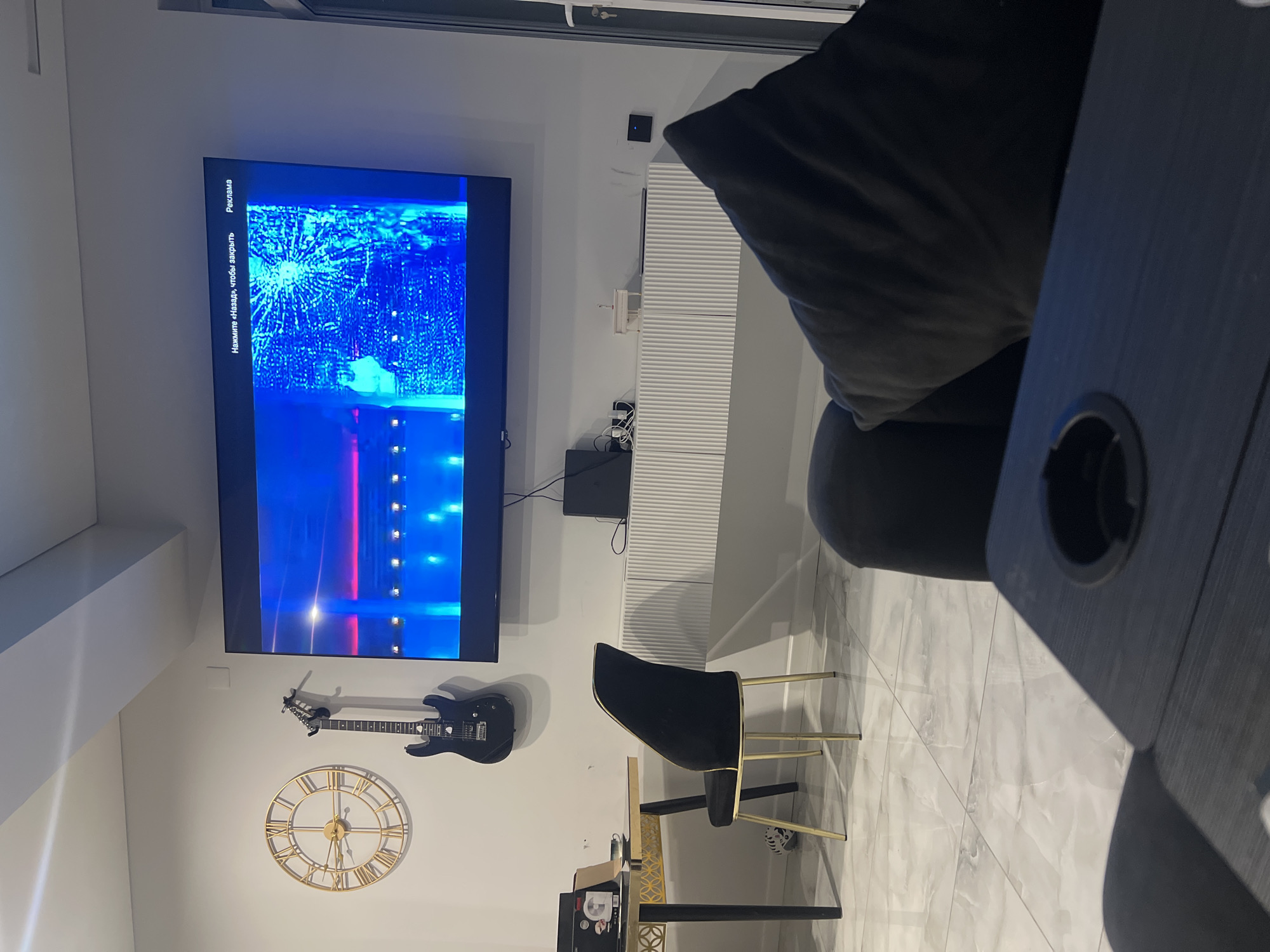 This image depicts a modern living room or entertainment space. The main subjects are a large flat-screen television mounted on the wall displaying a blue, abstract pattern with some red accents, and an electric guitar hanging on the wall below it. A large, round, gold-colored clock with Roman numerals is also visible on the wall.
In the foreground, a dark-colored armchair with gold legs is positioned near a desk. The desk has a black top and a patterned golden base. The floor is tiled with large, light-colored marble-effect tiles. Part of a dark sofa or chair is visible in the upper right, suggesting a seating area.
The scene appears to be indoors, with ambient lighting. There are no people visible in the image. The time of day is not explicitly indicated, but the indoor setting suggests it could be any time.
There is visible text on the television screen in vertical orientation on the left side. It appears to be in Russian and reads "Нажмите «Назад», чтобы закрыть" which translates to "Press 'Back' to close," and "Реклама" which means "Advertisement."