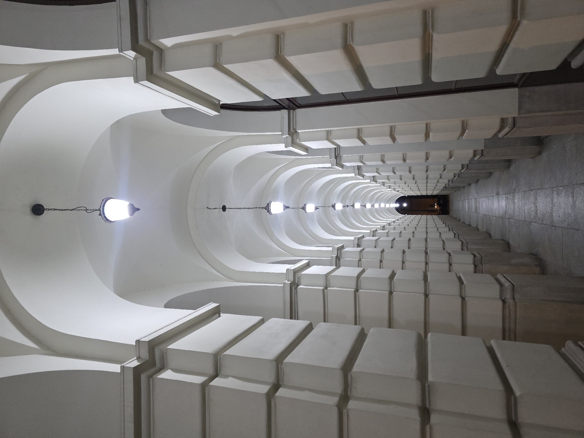 The image shows a long, white, arched corridor with rows of lights hanging from the ceiling. The corridor is lined with repeating architectural elements on both sides. The perspective creates a vanishing point at the far end, where a dark opening is visible. The scene is illuminated by the hanging lights, creating a sense of depth and symmetry. The corridor has white stone decorations and a dark strip in the middle of the top. This is the arcades on Zamkowy Square in Warsaw, Poland.