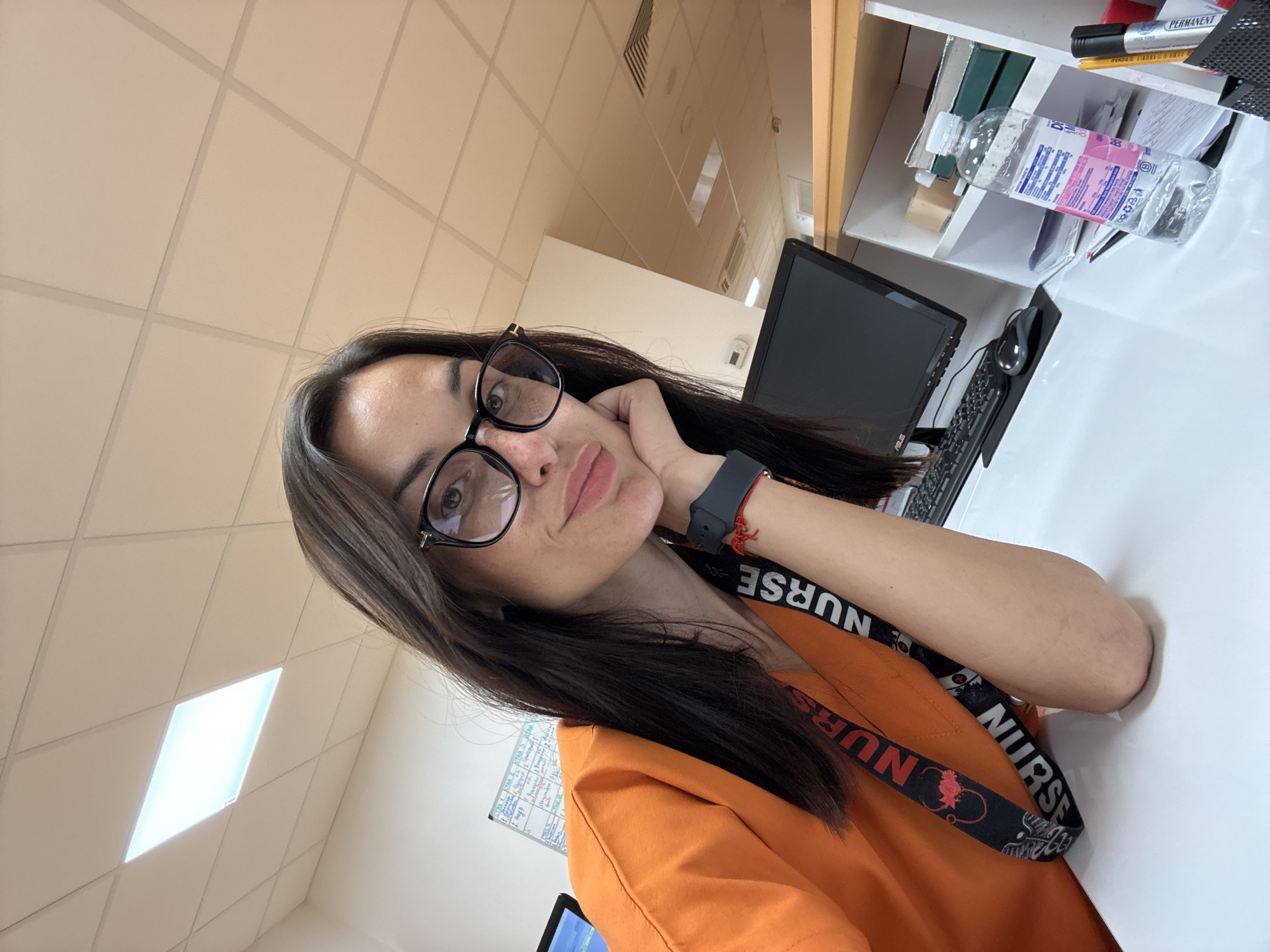 A young woman with dark hair and glasses is taking a selfie at a desk in what appears to be an office or hospital setting. She is wearing an orange scrub top and a lanyard with "NURSE" printed on it. Her right hand is resting on her chin, and she is looking towards the camera with a slight smile. On the desk in front of her is a computer monitor, keyboard, mouse, a bottle of water, and some papers. In the background, a ceiling with fluorescent lighting and a whiteboard with writing can be seen. The scene is brightly lit and appears to be indoors. The location is identified as Pleven, Bulgaria.