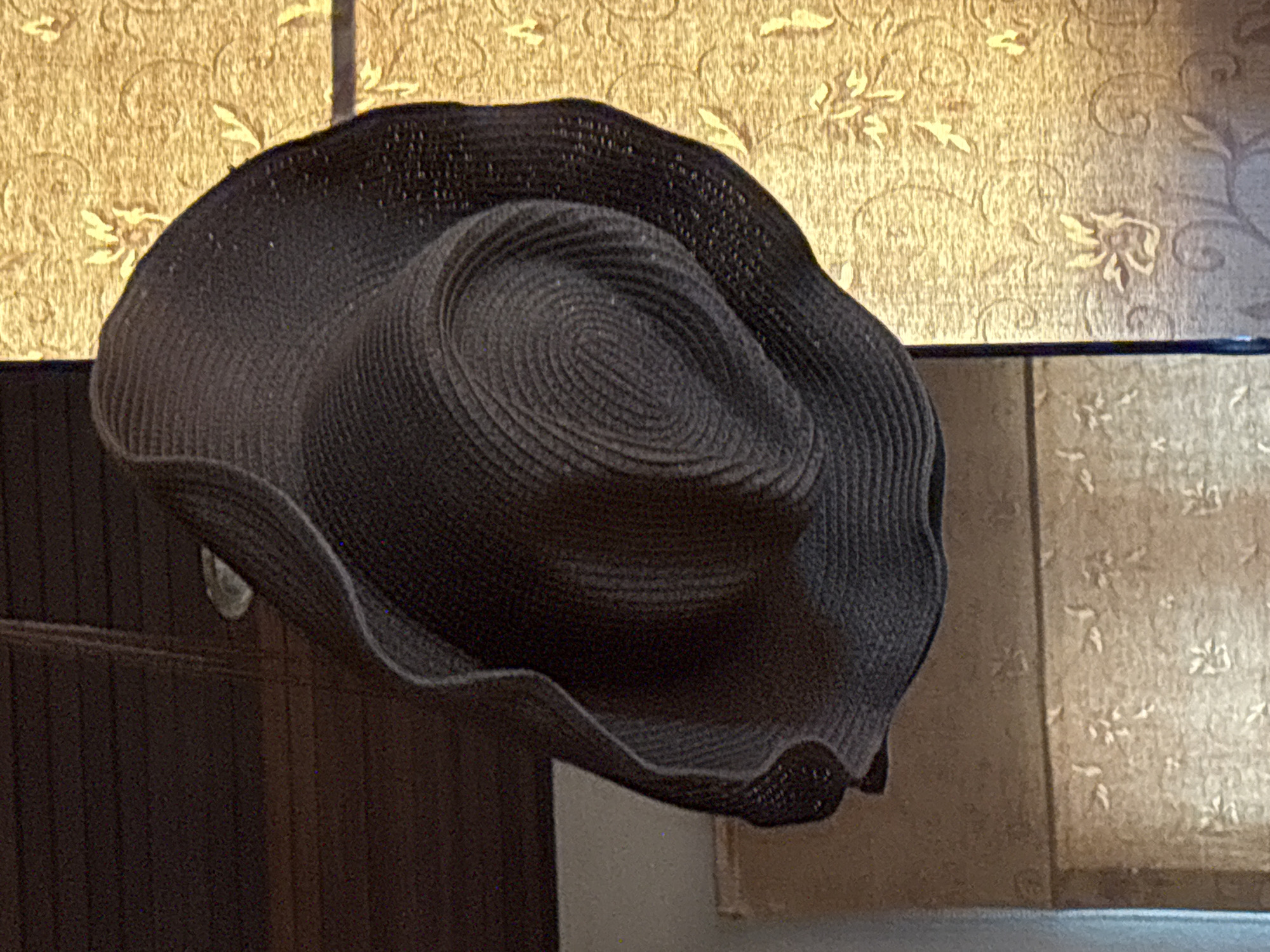 A dark-colored woven hat with a wide, undulating brim and a rounded crown rests on an unidentifiable object against a backdrop of textured wallpaper with a floral pattern. The location is บ้านบางรักษ์, Thailand.