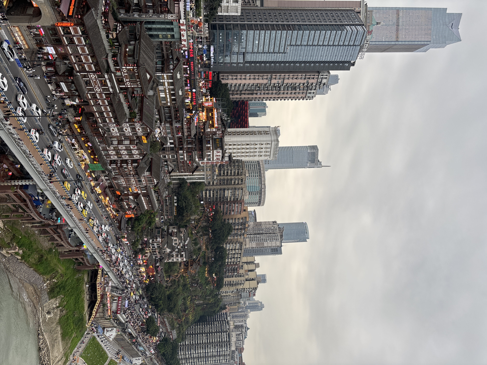 This is an elevated view in Yuzhong District, China, showcasing a blend of traditional architecture and modern skyscrapers. The bottom of the frame features a body of water with a shoreline transitioning to green vegetation. A roadway filled with vehicle traffic, primarily white taxis, bisects the frame. A bustling pedestrian area runs parallel, adjacent to traditional multi-story buildings with dark roofs and white facades. Beyond the traditional structures, modern high-rise buildings dominate the skyline, fading into an overcast sky.