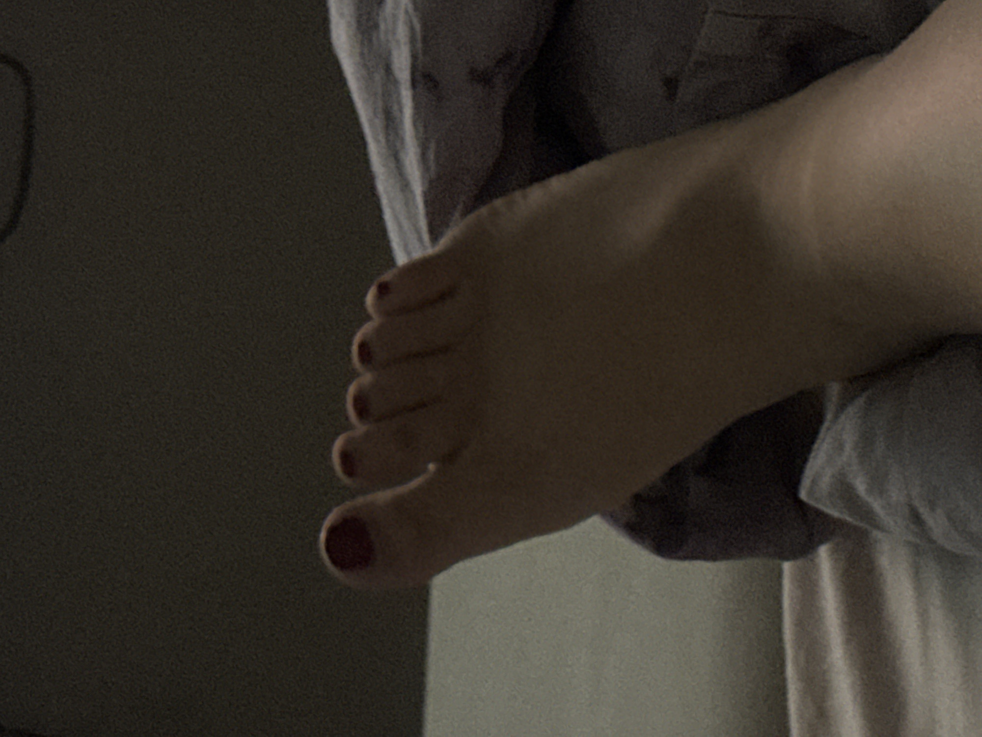 The image shows a close-up, low-light view of a person's left foot, resting on a bed with dark grey and lighter grey linens. The foot has red nail polish on all five toenails, with the big toe nail being the most prominent. The foot is angled, with the toes pointing upwards and slightly to the left. The background is dark and out of focus, suggesting an indoor setting. There is no indication of the specific location in Saratov, Russia.