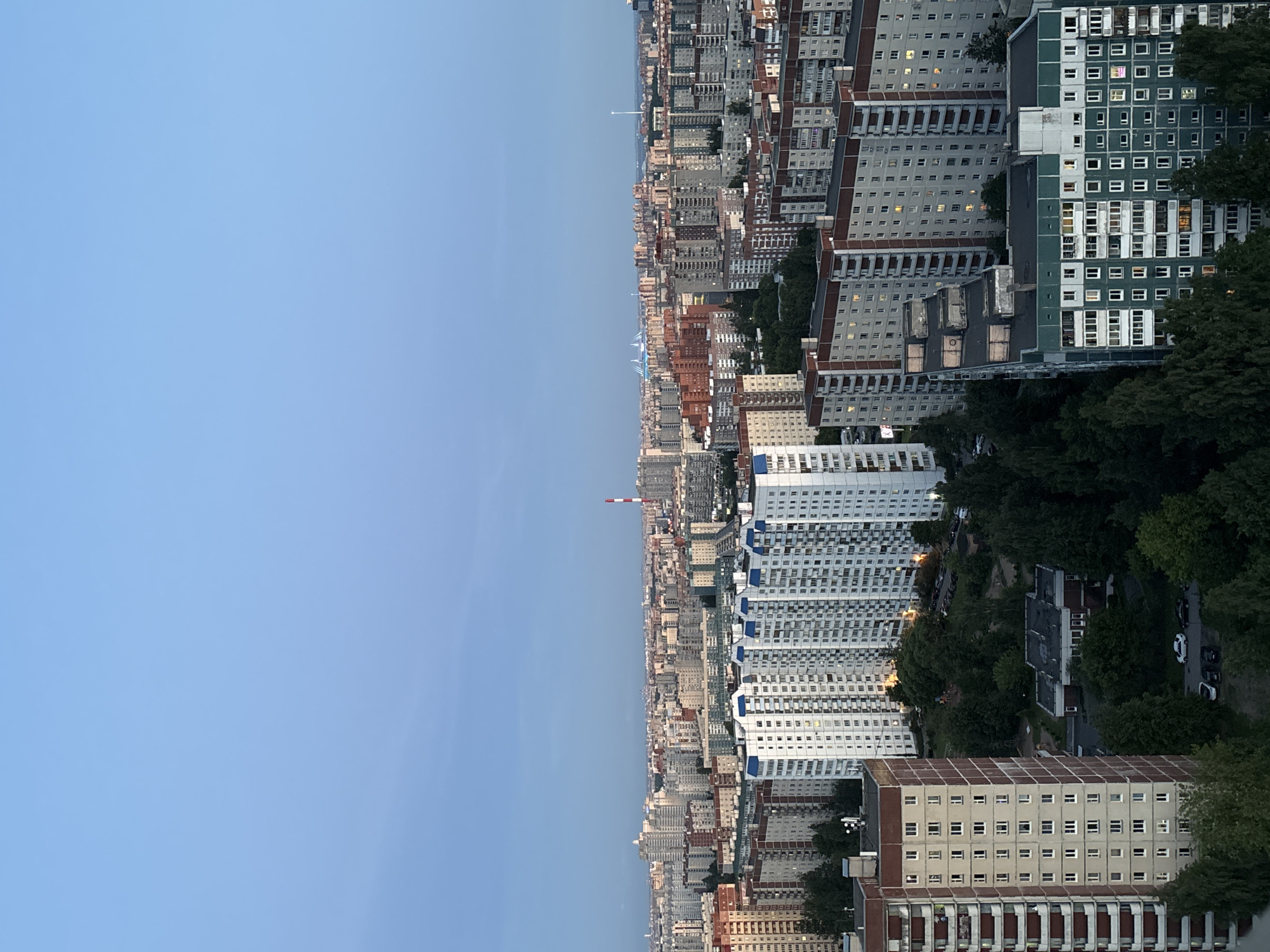 This is an aerial, full-frame, high-angle, outdoor, vertical, candid shot of a dense urban landscape at dusk. The scene is dominated by numerous apartment buildings of varying heights and architectural styles. The buildings are predominantly concrete, with many featuring light-colored facades and darker trim. Some buildings have balconies, while others have rows of windows. A few buildings have distinct features, such as a building with a green facade in the foreground on the right, and a tall, red and white striped smokestack in the mid-ground on the right.
The foreground on the left shows the side of a tall, light-colored apartment building with reddish-brown balconies. This building is bordered by dark green trees. In the mid-ground, a cluster of tall, white apartment buildings with blue accents stand out. These buildings are interspersed with dense green trees. Further into the background, a sprawling cityscape of more apartment buildings stretches to the horizon, with hints of distant industrial structures and possibly bridges or other large infrastructure visible in the far distance under a pale blue, cloudless sky that transitions to a lighter hue near the horizon. The lighting suggests the late evening or early morning, with some windows in the buildings illuminated from within. The overall impression is of a large, residential urban area during a quiet time of day.