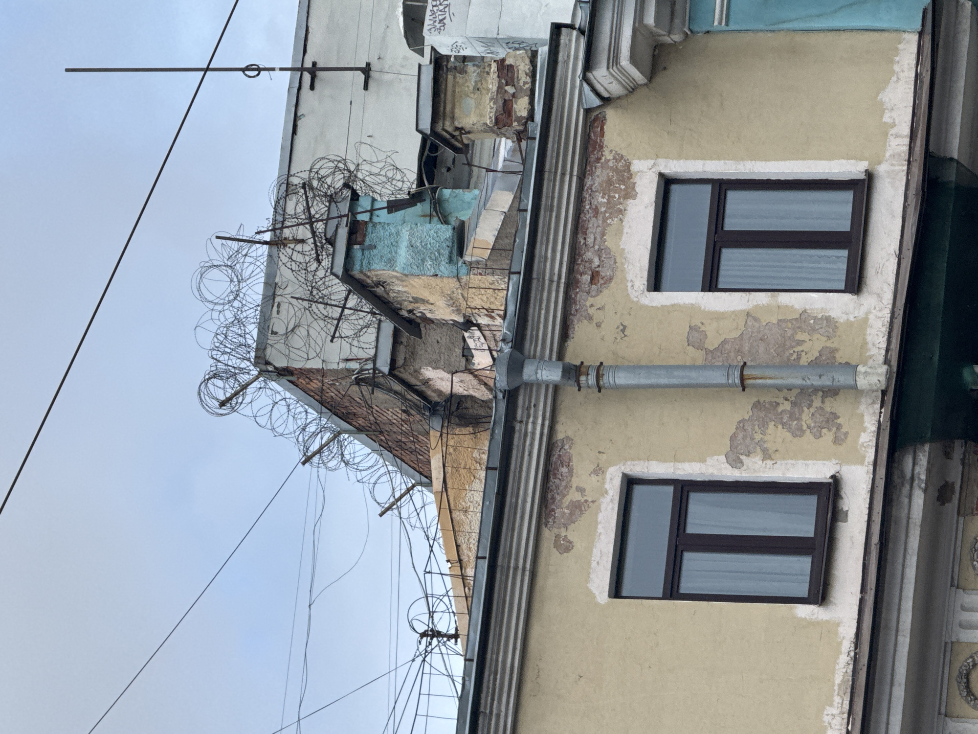 The image shows a building with two windows on its facade. The building has a distressed yellow paint finish with areas of exposed brickwork. A metal downspout runs vertically along the side of the building. Above the facade, on the rooftop, there are various structures including chimneys and barbed wire fencing. The sky in the background is overcast. The scene is likely in Saint Petersburg, Russia, given the architectural style.