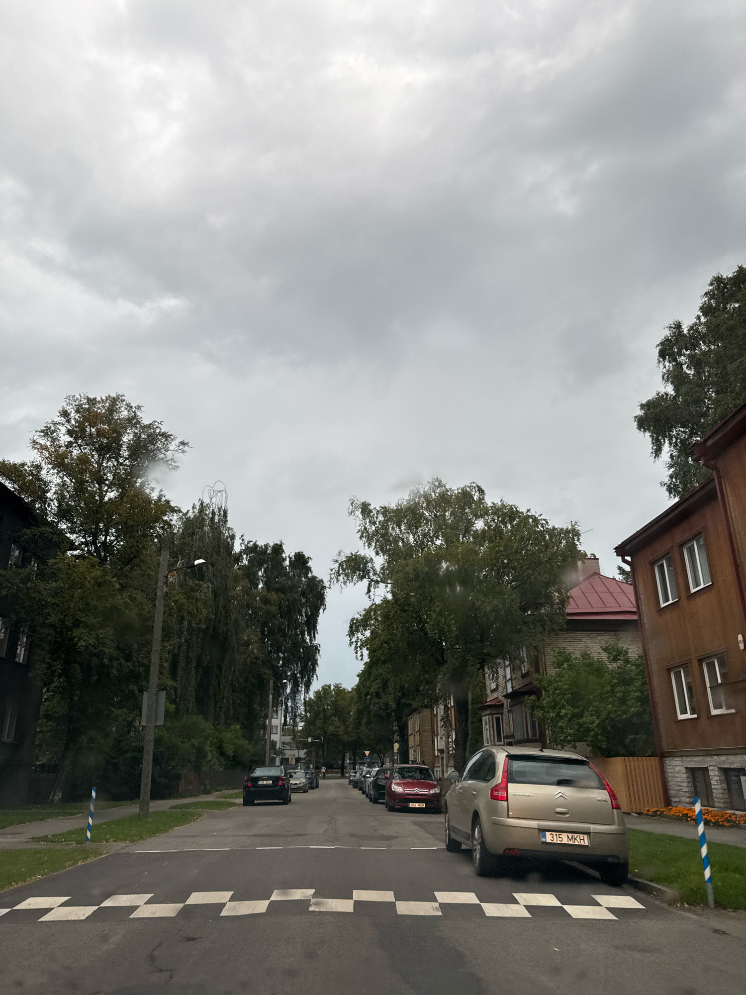 The image depicts a street scene in Tallinn, Estonia, under a cloudy sky. A checkered pedestrian crossing is visible in the foreground. Several cars are parked and driving on the asphalt road. On the right side of the street, there is a wooden building with white-framed windows and a red-tiled roof. The left side of the street features buildings and dense trees. Streetlights and trees line the road, contributing to a residential or suburban atmosphere.