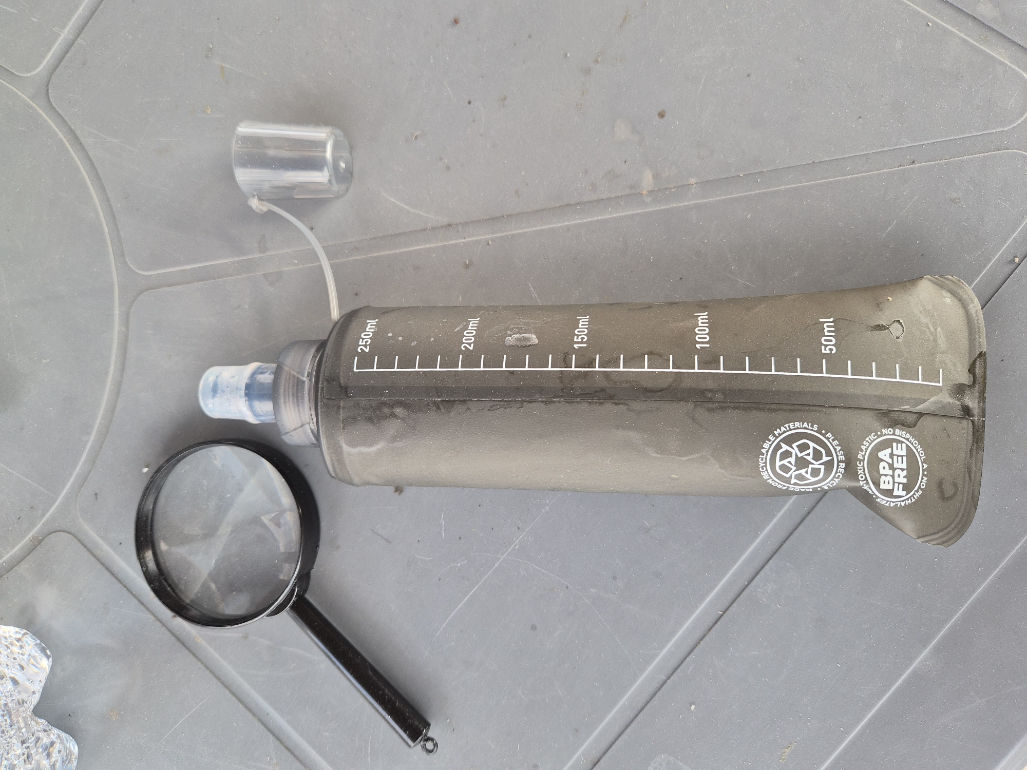 A soft-sided, black water bottle with volume markings in white is laid out on a gray, textured surface, possibly a table or plastic container lid. The bottle has a clear plastic cap attached by a thin white cord. A black-handled magnifying glass rests to the left of the bottle, and a small piece of clear, textured material is partially visible in the bottom left corner. The water bottle features markings indicating volumes of 50ml, 100ml, 150ml, 200ml, and 250ml. Circular text and symbols are printed on the right side of the bottle, including "RECYCLABLE MATERIALS," "PLEASE RECYCLE," "MADE FROM RECYCLED," "NON-TOXIC PLASTIC," "BPA FREE," and "NO BISPHENOL A. NO PHTHALATES." The scene appears to be outdoors, possibly in daylight, given the lighting and the texture of the surface. The overall impression is one of an outdoor or camping setting, with an emphasis on practical, reusable items.