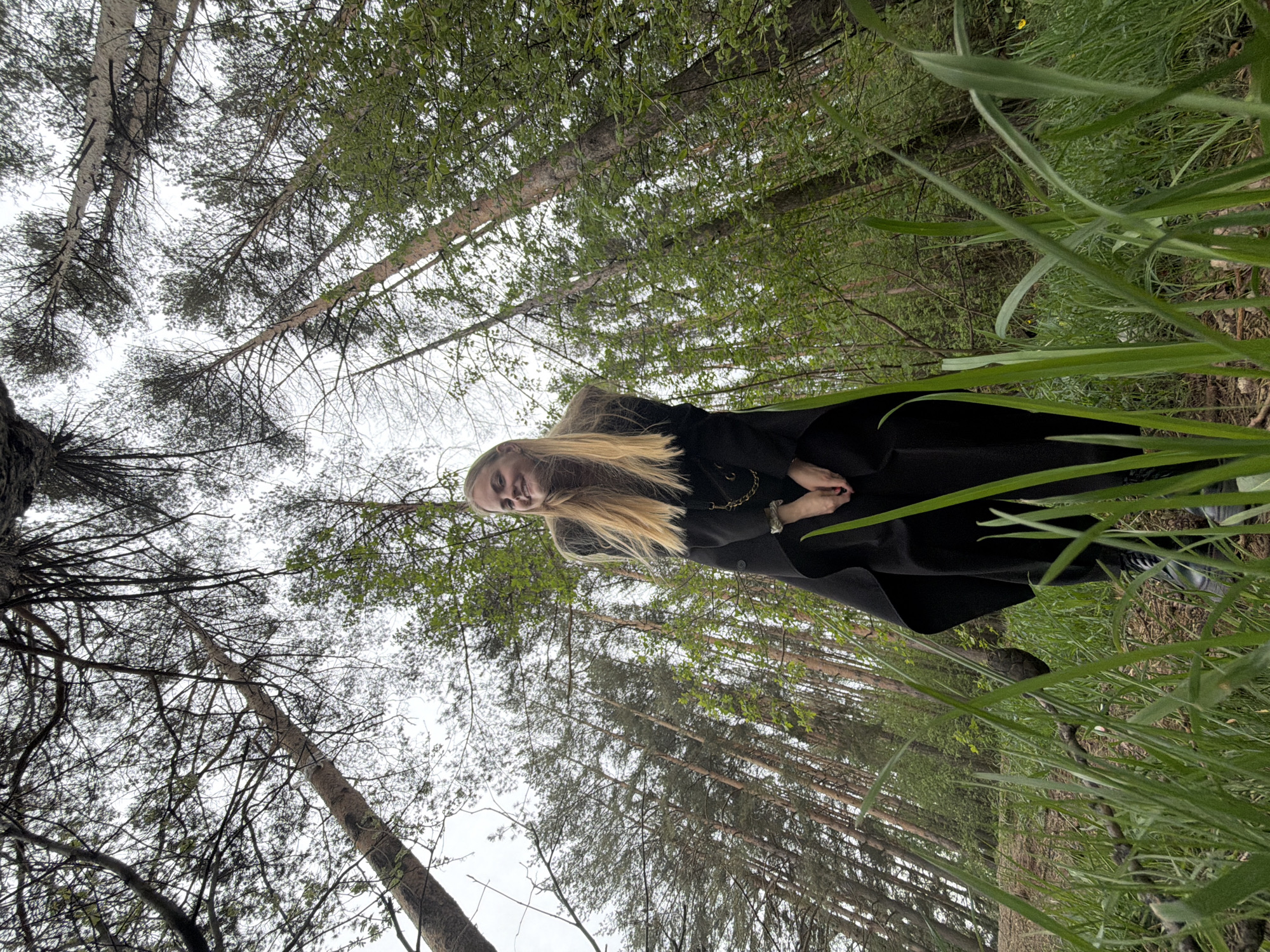 A woman stands in a forest. She has long blonde hair and is wearing a black coat. She is surrounded by tall trees and green vegetation. The location is Izhevsk, Russia.