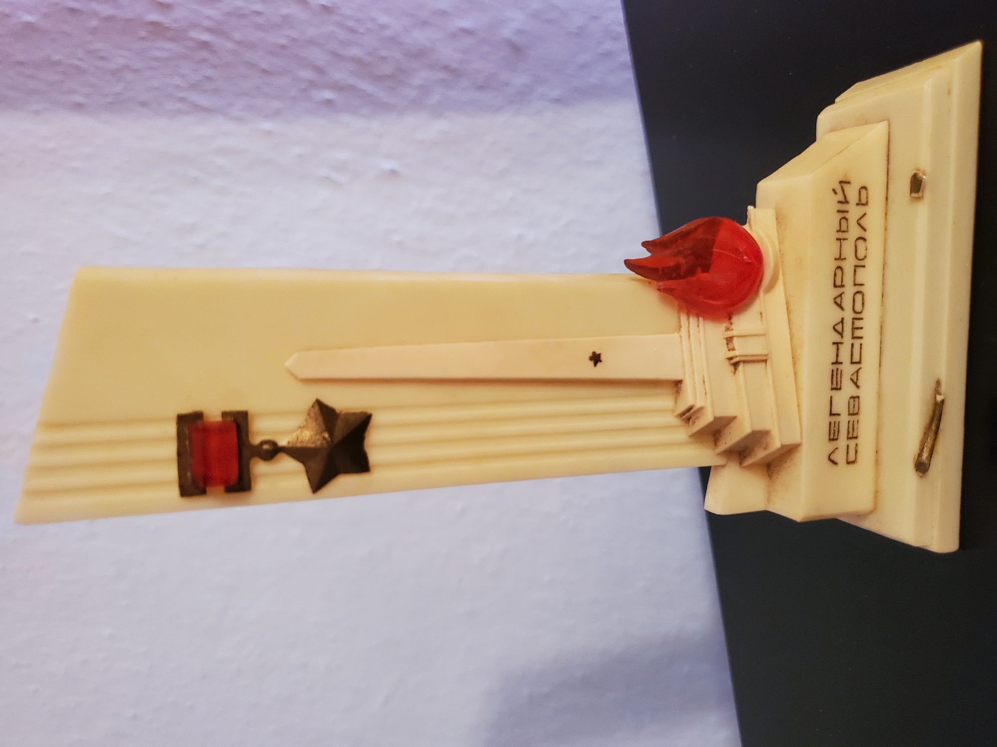 A cream-colored, rectangular statuette depicts a monument with several symbolic elements. At the top, a dark metal star is suspended from a red ribbon, which is attached to a small dark metal bar. Below this, a tall, slender obelisk rises from a tiered base. To the right of the obelisk, a small, red, flame-shaped ornament is affixed. The base of the monument is inscribed with Cyrillic text that reads "ЛЕГЕНДАРНЫЙ СЕВАСТОПОЛЬ" (Legendary Sevastopol). A small, dark metallic object, possibly a bullet or casing, is embedded in the front of the base. The statuette is positioned on a dark, flat surface, with a textured, light-colored wall visible in the background. The lighting casts subtle shadows, highlighting the relief details of the statuette. The image is taken from a slightly low angle, focusing on the central object.