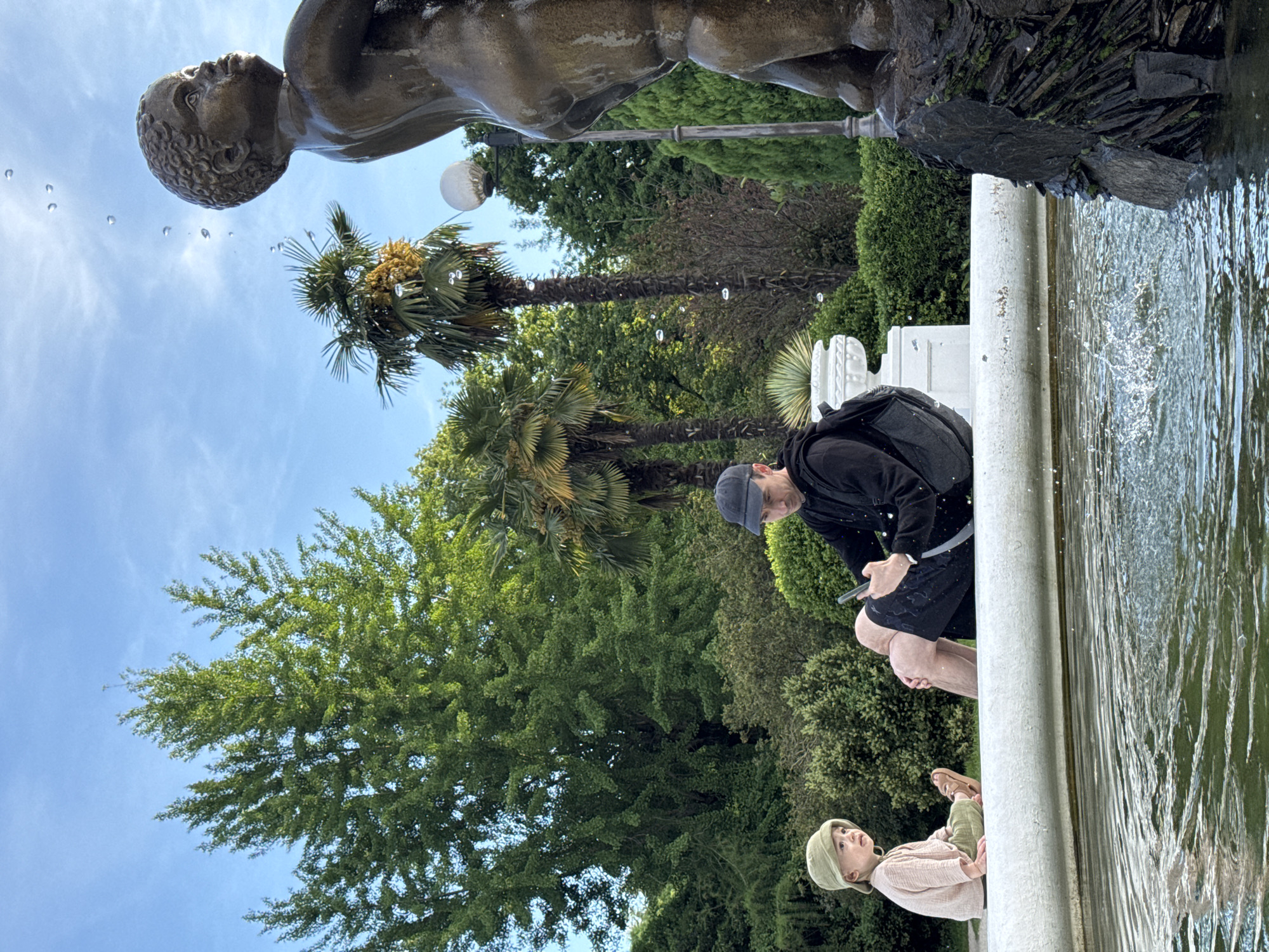 The image depicts a park scene in Sirius, Russia, featuring a fountain. An adult male wearing a black jacket and blue baseball cap sits on the edge of the fountain, using a mobile device. A child wearing a beige shirt and a green hat sits to the left of the man, also on the edge of the fountain. Water is flowing over the edge of the fountain into a pool. A bronze statue of a male figure stands atop the fountain. Several trees and palm trees are visible in the background under a blue sky.