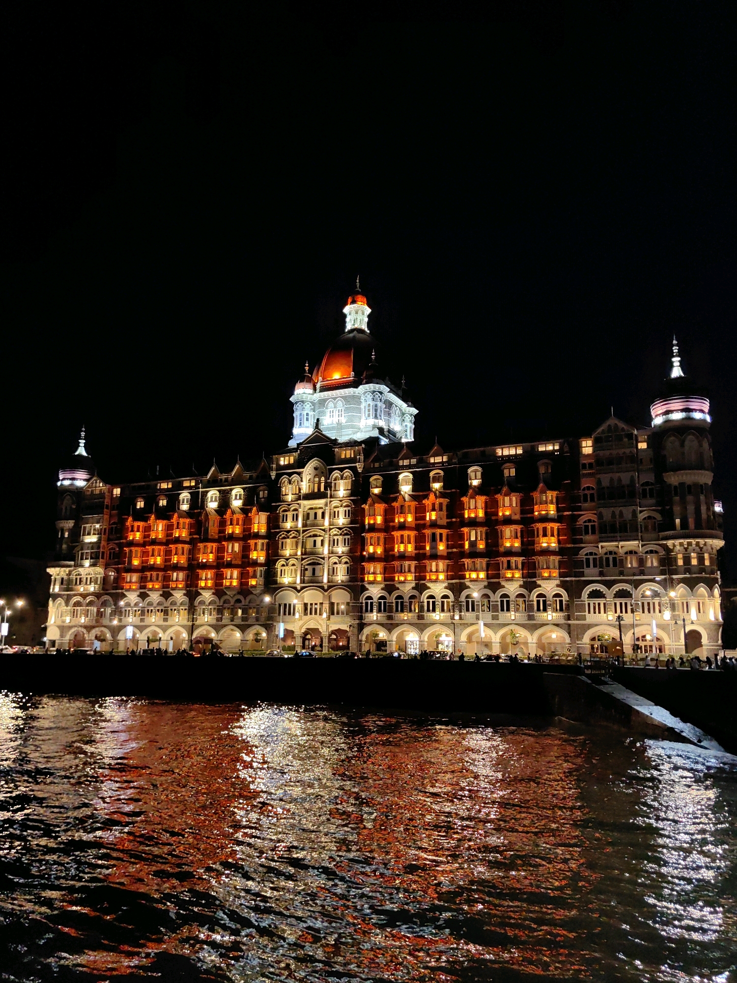 The image depicts the Taj Mahal Palace Hotel, a historic luxury hotel in Mumbai, India, at night. The grand building is illuminated, with portions of its facade lit in warm orange and yellow hues. A prominent red dome caps the central structure, and two smaller domes are visible on either side. The hotel is situated along the waterfront, and the lights from the building are reflected in the dark water in the foreground. The sky is black, indicating nighttime. A sidewalk or promenade with scattered figures of people and vehicles is visible along the base of the hotel. The reflection in the water is vibrant, showing streaks of orange, yellow, and white light.