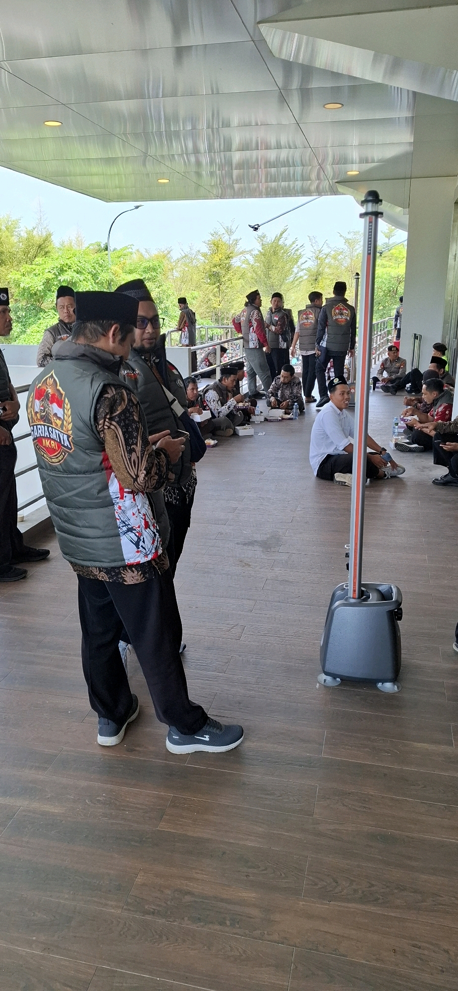 In the city of Blulukan, Indonesia, a group of men are gathered outdoors under a modern, reflective canopy with recessed lighting. The ground is paved with wooden planks. In the foreground, two men stand and converse. The man on the left wears a dark vest with a circular emblem, a patterned long-sleeved shirt, and black trousers. The man on the right, wearing glasses and a similar vest and shirt, looks at a mobile phone. Behind them, other men are seated on the ground, some eating from containers, while others stand and observe. One man is seated apart on the right, wearing a light blue shirt and black trousers. A tall, dark grey portable device with a red and white stripe is positioned near the center right of the frame. Trees and foliage are visible in the background.