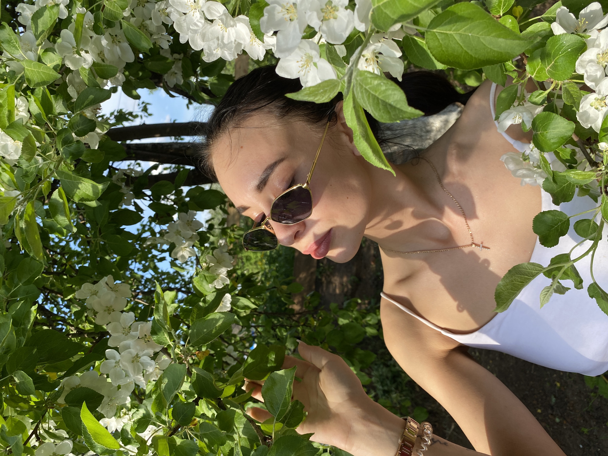 A young woman with dark hair pulled back, wearing sunglasses and a white top, is positioned amidst flowering branches with white blossoms and green leaves. She is touching a leaf with her right hand, on which she wears a gold watch. A gold necklace adorns her neck. The background shows a glimpse of the sky and tree branches. The location is in a garden or orchard-like setting in the city of Chaikovsky, Russia.