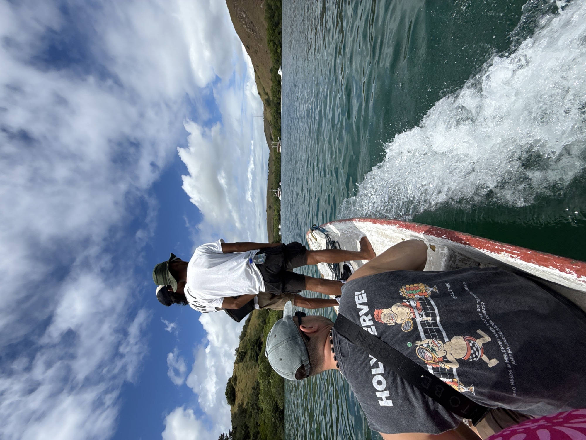 The image shows people in a small boat on a body of water, possibly a lake or bay. The boat is creating a wake, indicating movement. The scene includes a mountainous or hilly landmass in the background with green vegetation. The sky has scattered clouds. The location is indicated as being in Unknown, Indonesia. The image is rotated from a normal orientation, which may affect perception of the composition.