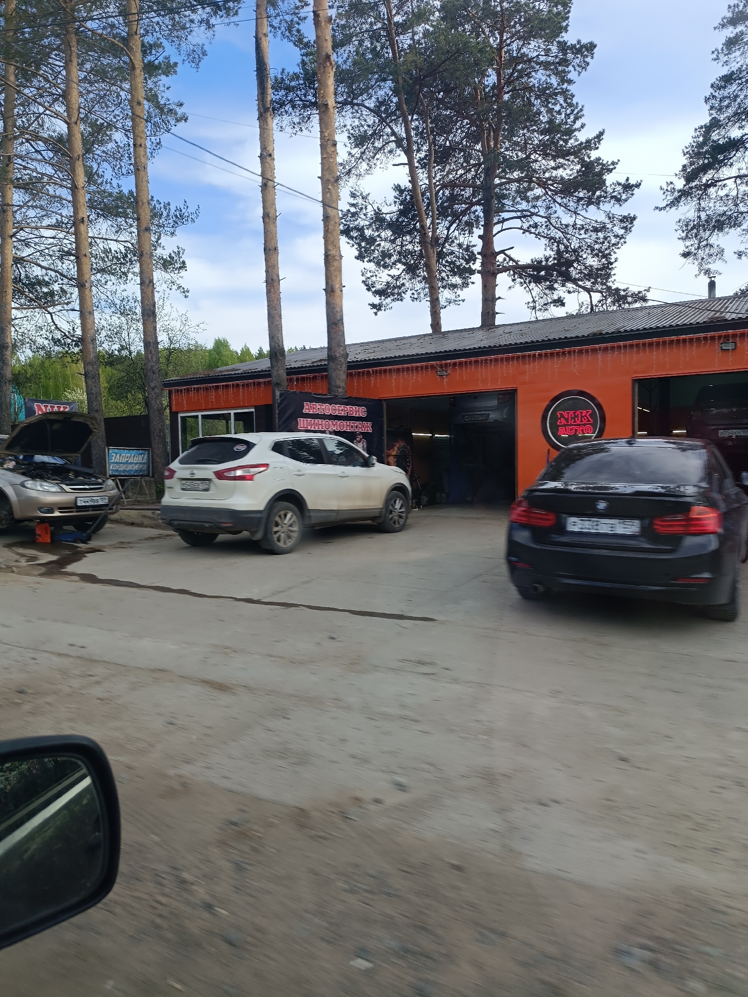 The image shows a roadside scene in what appears to be a commercial or industrial area of the city of Chaikovsky, Russia. The main focus is a single-story, orange building with multiple garage-style doors. Signage in Russian above the garage bays indicates automotive services, potentially including tire repair ("шиномонтаж"). There is a large round sign with the text "NHK AUTO".
Three vehicles are visible. At the left of the frame, a gray car has its hood open, and a jack is positioned underneath. A white SUV is parked in front of the central garage door. A black sedan is pulling into the rightmost garage bay.
The area in front of the building is a gray, unpaved surface. Tall trees surround the building, and glimpses of other buildings and green foliage are visible in the background. Part of a car mirror is in the lower-left corner.