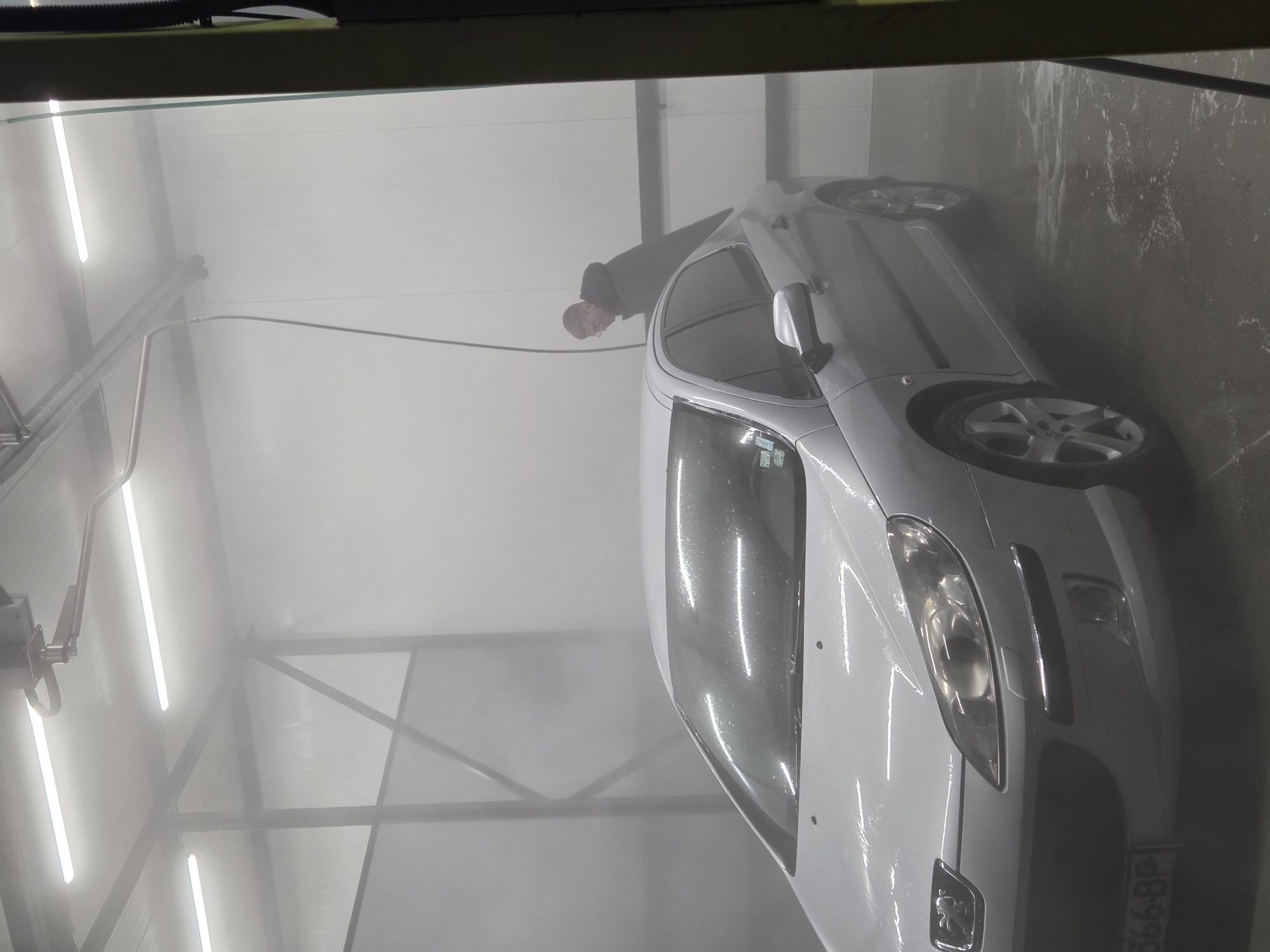 A white car is being washed inside a car wash. A man in a dark jacket is near the car's roof. Fluorescent lights illuminate the interior. A hose extends from the left side of the image toward the car. Part of a license plate with the letters "BP" is visible. The city location is not discernible from the image.