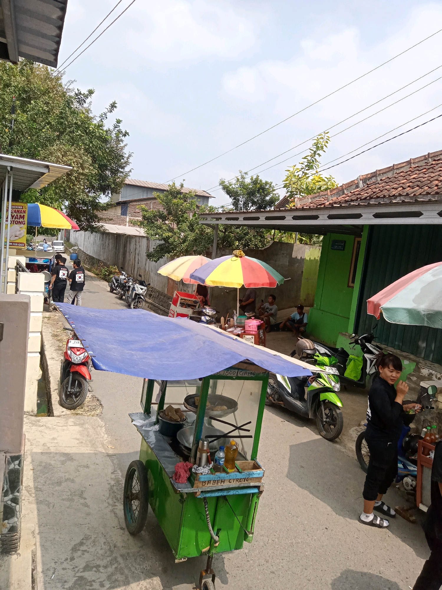The image depicts a street scene in Gumiwang, Indonesia, with a focus on a food cart. The street is paved with concrete and appears to be a relatively narrow road. On the left side of the image, there is a building with a metal roof and a white wall with decorative elements. Further down the street, more buildings with tiled roofs are visible, and lush green trees line the area.
The main subject is a bright green food cart with a blue tarp covering its top, providing shade. The cart has wheels and contains various items, including a pot or bowl, bottles of condiments, and what appear to be food items. A red motorcycle is parked to the left of the cart.
Several people are present. Two individuals wearing black t-shirts with a white emblem are standing near the left side of the image, possibly interacting with a vendor or waiting. Another person, a woman with dark hair, is standing to the right of the food cart, wearing black clothing and white socks with sandals. She appears to be holding something in her hand and looking to the right. Several motorcycles are parked along the street.
Colorful umbrellas, red, yellow, and blue, are spread out over some of the stalls and areas, indicating a marketplace or food vendor area. The sky is partly cloudy and bright, suggesting it is daytime. The overall atmosphere is that of a casual, outdoor commercial setting.