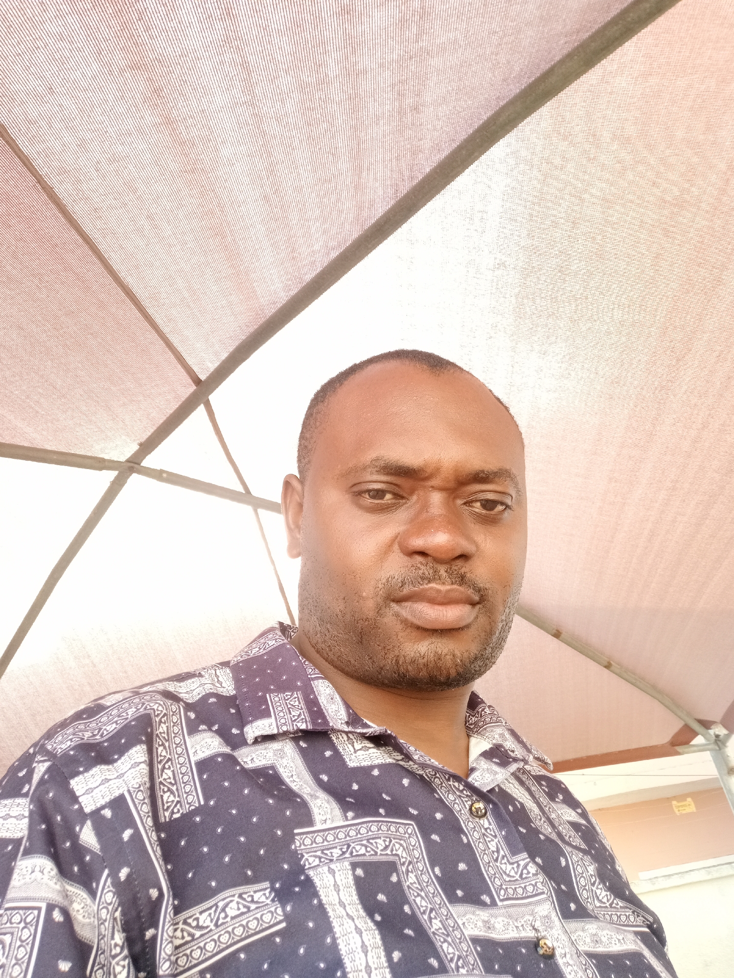 A medium shot depicts a dark-skinned man in Gwarinpa, Nigeria, under a sunshade structure with a striped, tent-like fabric roof supported by metal beams. He wears a navy blue shirt with a white patterned design and has a dark, short haircut with closely trimmed facial hair. The background beyond the sunshade structure shows parts of a building with a beige exterior wall and a small, yellow sign. The man is looking directly at the camera with a slight frown.