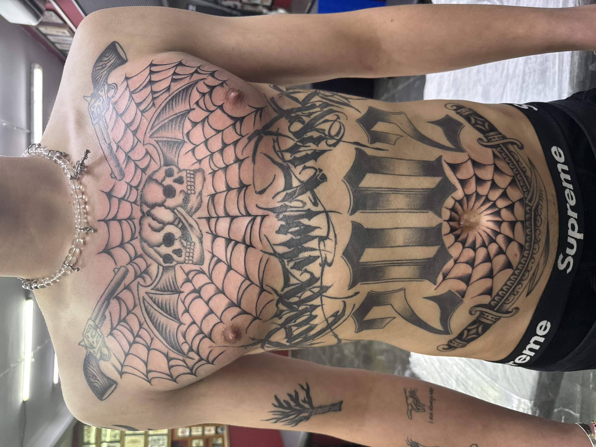 A person with extensive tattoos is shown from the chest down, wearing black Supreme underwear. The tattoos include a web-like design with skulls and guns on the chest, a large tattoo across the abdomen that appears to incorporate lettering and a web-like pattern around the navel, and a dagger on the side of the abdomen. There are also tattoos on the arms. A beaded necklace is around the person's neck. The background is blurred and indistinct. The location of this image is unknown.