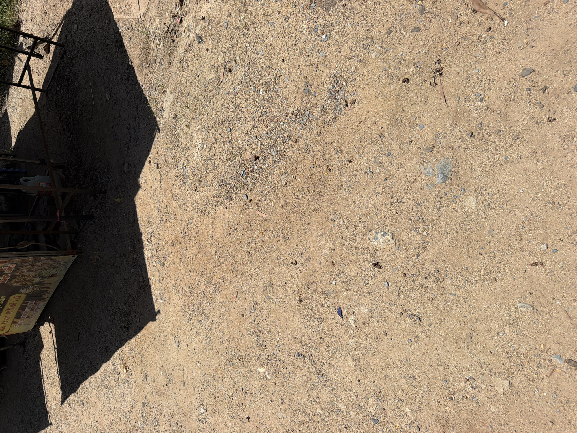The image depicts a sandy, gravelly ground surface in an outdoor environment, likely in Baan Bo Phut, Thailand, based on the contextual information. A dark shadow cast by an overhead structure, possibly a market stall or a temporary shelter, dominates the upper left and central portions of the frame. The ground is uneven, with visible pebbles, small stones, and patches of dry earth. Scattered across the surface are a few fallen leaves, some small pieces of debris, and what appear to be tiny dark droppings or soil clumps. In the upper portion of the image, the edge of a stall structure is visible, including a metal frame and a colorful banner with text in an Asian script, suggesting a commercial or food vending area. The lighting indicates it is daytime, with strong sunlight creating sharp shadows. There are no people or animals clearly visible in the image, focusing instead on the texture and composition of the ground and the surrounding rudimentary structures.
