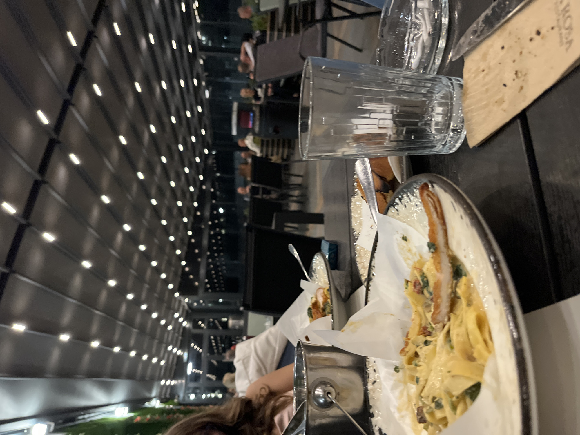 The image shows a table setting at a restaurant, likely outdoors given the view of a building facade with numerous lights visible in the background. A glass of water and silverware are placed on the table, alongside a partially consumed plate of pasta with chicken or other protein. People are visible at other tables in the background, suggesting a populated dining area. It is not possible to confirm the location in Karlovo, Bulgaria based on visual elements alone.