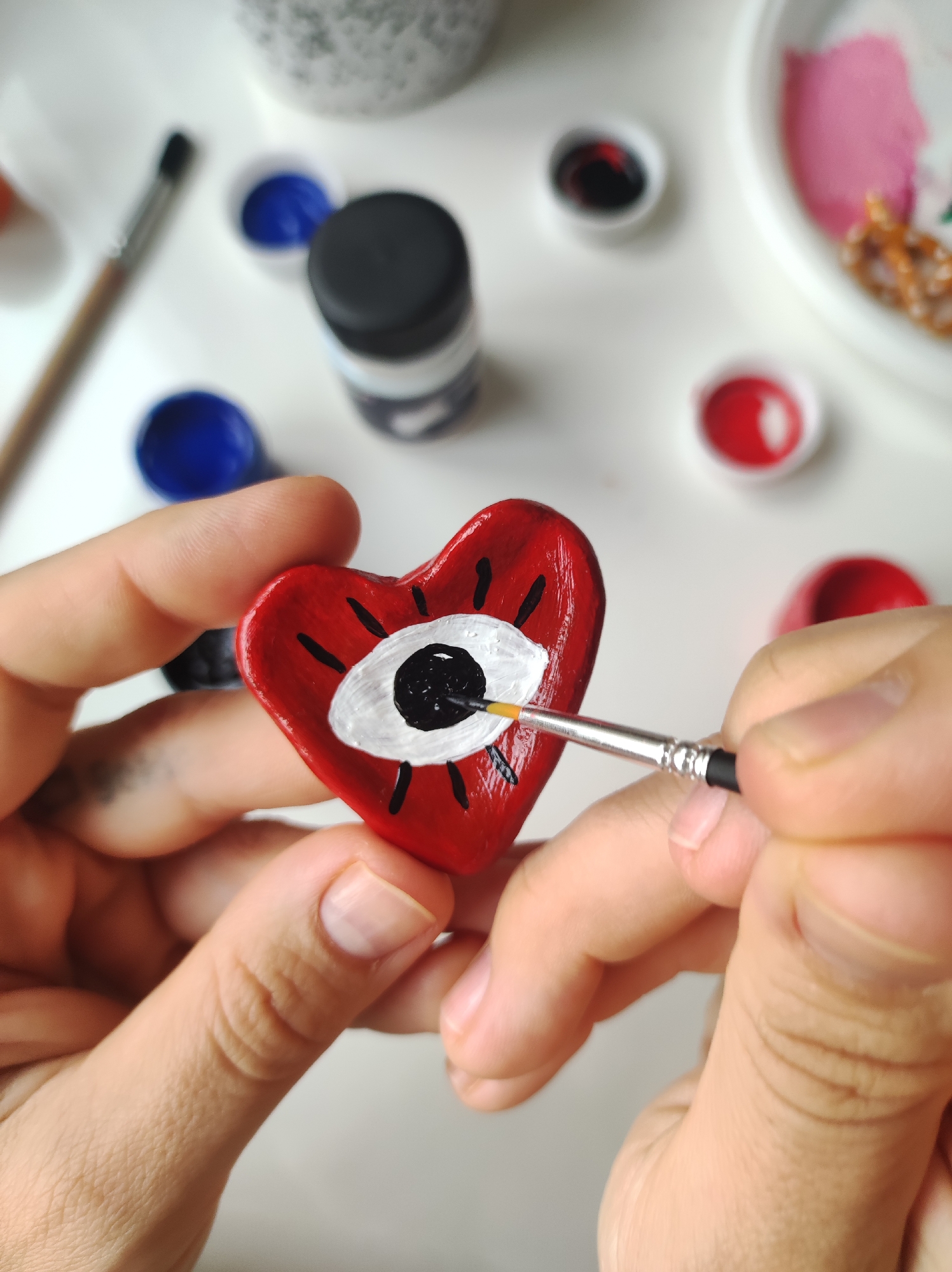 A person is painting a small, red heart-shaped object with a white and black eye design. The person is holding the heart in one hand and using a thin paintbrush to apply black paint to the eye. Several small containers of paint in various colors (blue, red, black, white, pink) and a paintbrush are visible on a white surface in the background. The scene appears to be indoors, possibly a table where crafting is taking place. There is nothing in the image to indicate it's in Chaikovsky, Russia.