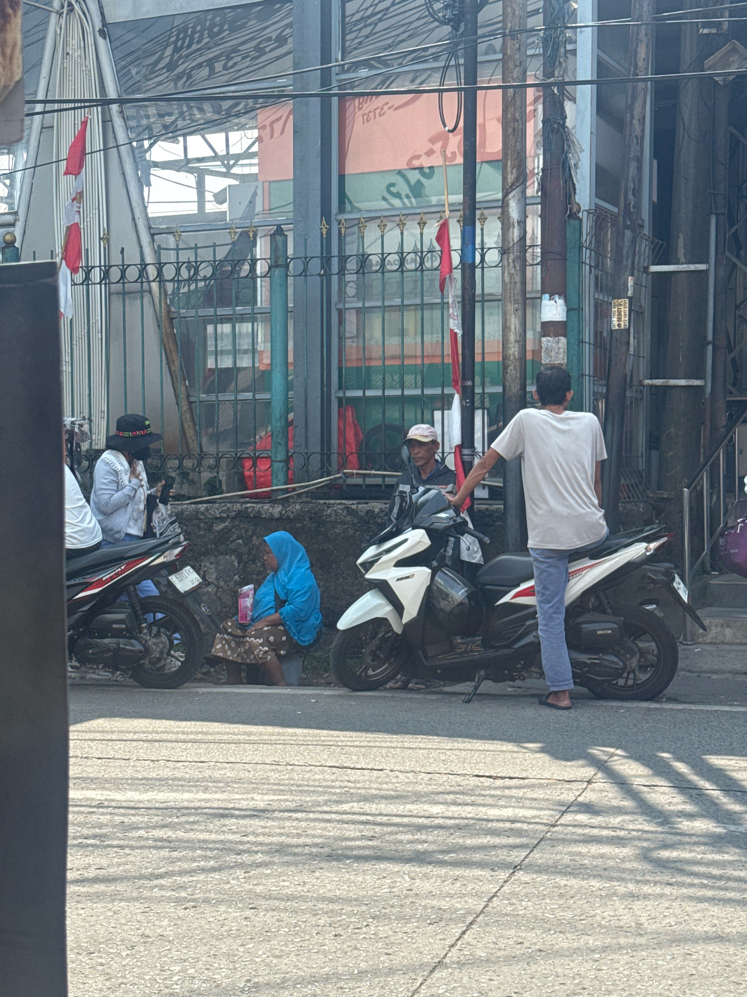 In Sukaraja, Indonesia, on a sun-dappled street, a group of individuals are gathered near parked motorcycles. A woman wearing a blue hijab and patterned skirt sits on the ground, holding a pink packet, while a man in a white t-shirt and blue jeans stands beside a white motorcycle. Another man in a dark jacket and a light-colored cap is seated on a black motorcycle. In the background, a black fence with circular designs separates the sidewalk from a building with large windows and an orange facade displaying text. Indonesian flags are visible, attached to poles along the fence. The scene is set against a backdrop of power lines and urban structures.
