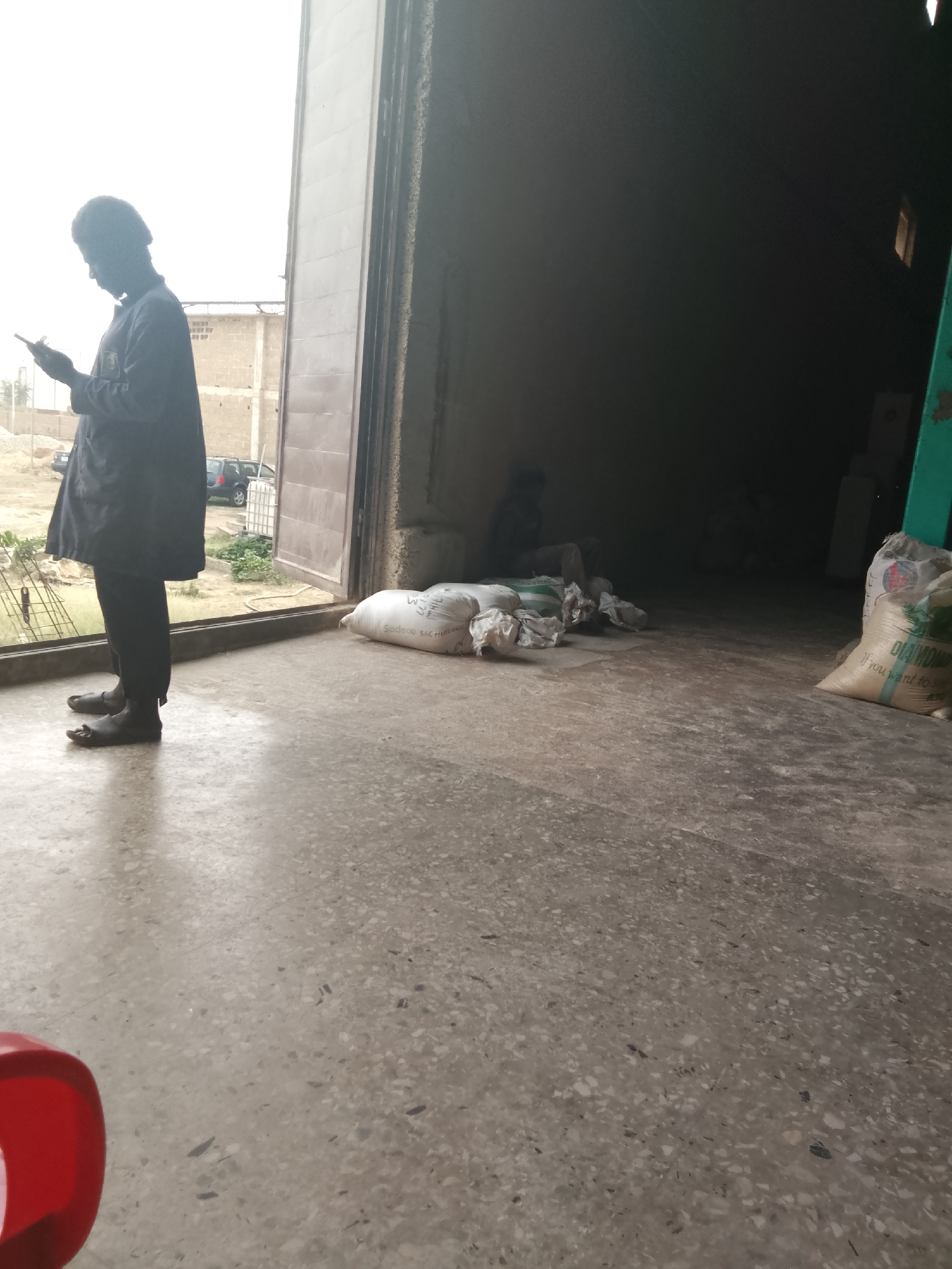 The image is taken from inside a large building, looking out through a wide, open entrance into a bright outdoor environment. The floor inside is a light grey, speckled concrete or terrazzo. A red object, possibly a bucket, is partially visible in the bottom left foreground.
On the left side of the open doorway, a person stands in profile, silhouetted against the bright exterior light, deeply engaged with their mobile phone. They are wearing a dark, long-sleeved garment and dark trousers, with dark open-toed sandals on their feet.
Inside the building, which is largely in shadow, several large sacks are placed on the floor near the entrance. One white sack clearly displays the text "SODECO SAC HUILE". Deeper within the dark interior, another heavily shadowed person is vaguely visible, sitting or kneeling on the floor. On the far right, another large, light-colored sack is visible, bearing the text "DIAMOND" and partially legible words, "If you want to s...".
The outdoor scene, seen through the doorway, features an unpaved or dirt area with some sparse greenery. In the background, there are buildings constructed with cinder blocks, a dark-colored car, and a white plastic container or tank. The strong, bright light outside indicates it is daytime, likely a sunny day. The scene depicts a mundane moment, possibly at a warehouse or storage facility in Jos, Nigeria, with the person on the phone engaged in a common modern activity amidst an industrial or commercial setting.