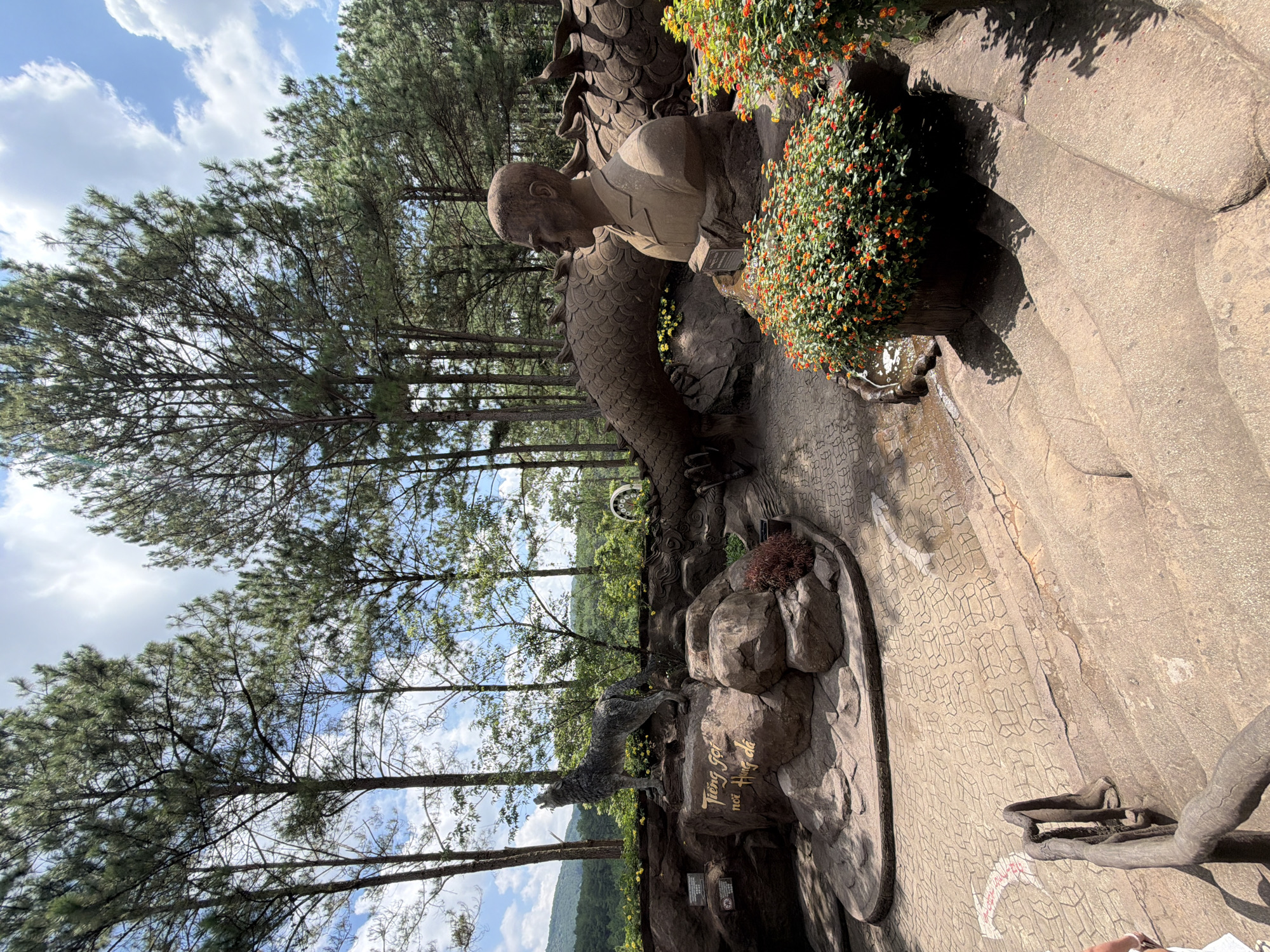 The image depicts a scenic outdoor sculpture garden in Da Lat, Vietnam, likely within the Datanla Waterfall area. The foreground features a set of wide, stone steps with a rustic, tree-like railing on the left. The steps lead up to a paved area adorned with intricate patterned stonework.
Dominating the midground is a large, intricately sculpted dragon, its textured body winding through the scene. Resting on a rock formation to the left of the dragon is a statue of a wolf, head tilted back as if howling. Inscribed on the rock beneath the wolf are golden Vietnamese characters that translate to "Call of the Mountains and Rivers."
To the right of the dragon's body, a bust of a man in formal attire is prominently displayed, facing left. This sculpture is framed by vibrant clusters of small, orange and red flowers. The background is filled with tall pine trees, characteristic of Da Lat's highlands, and a hazy, distant mountain landscape under a partly cloudy sky. The overall impression is one of a carefully curated cultural and natural attraction.