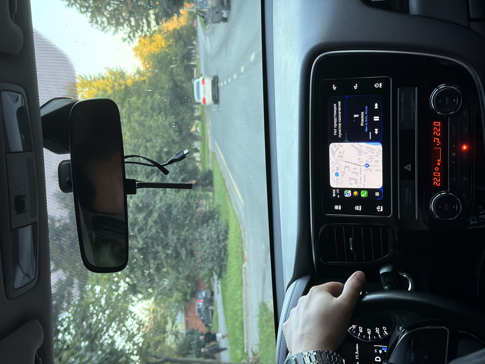 The image shows the interior of a car driving through Kazan, Russia. The driver's hand is on the steering wheel, and the dashboard displays a navigation map and music controls. The road is lined with trees and houses, suggesting a suburban or residential area. The sky is clear, indicating fair weather.