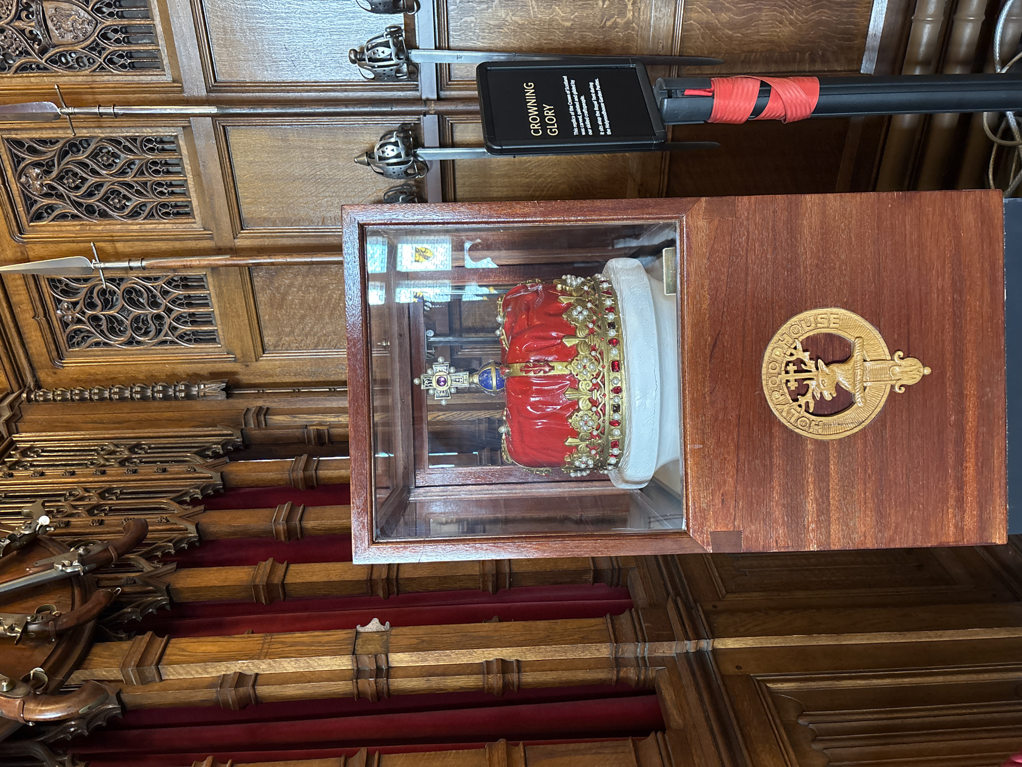 The image shows a display case containing a replica of the Crown of Scotland, located in the City of Edinburgh, United Kingdom. The replica is a red and gold crown adorned with jewels, sitting atop a white stand within the glass case. To the right of the case is a wooden display board with a gold emblem of the Holyroodhouse logo. Above and behind the case are historic weapons, including spears and swords, mounted on a carved wooden wall. A black sign with red tape indicates "Crowning Glory," detailing that the replica was carved, painted, and gilded, and is used during the Holyroodhouse Garden Parties. The surrounding environment includes intricately carved wooden paneling and red fabric-covered beams, suggesting a historic or ceremonial setting.