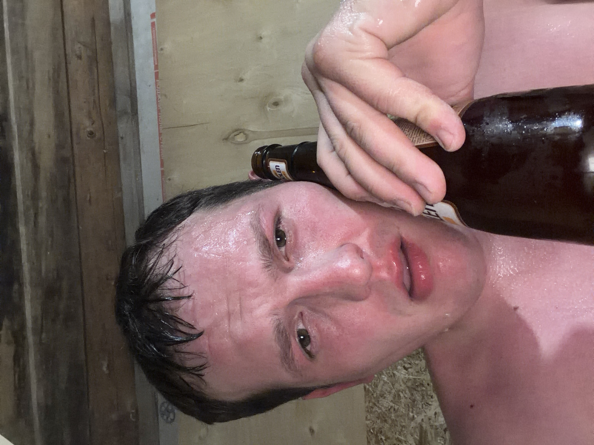 A close-up, slightly tilted shot shows a perspiring man holding a brown beer bottle against his head. The background includes wooden planks or paneling. The location is unknown.