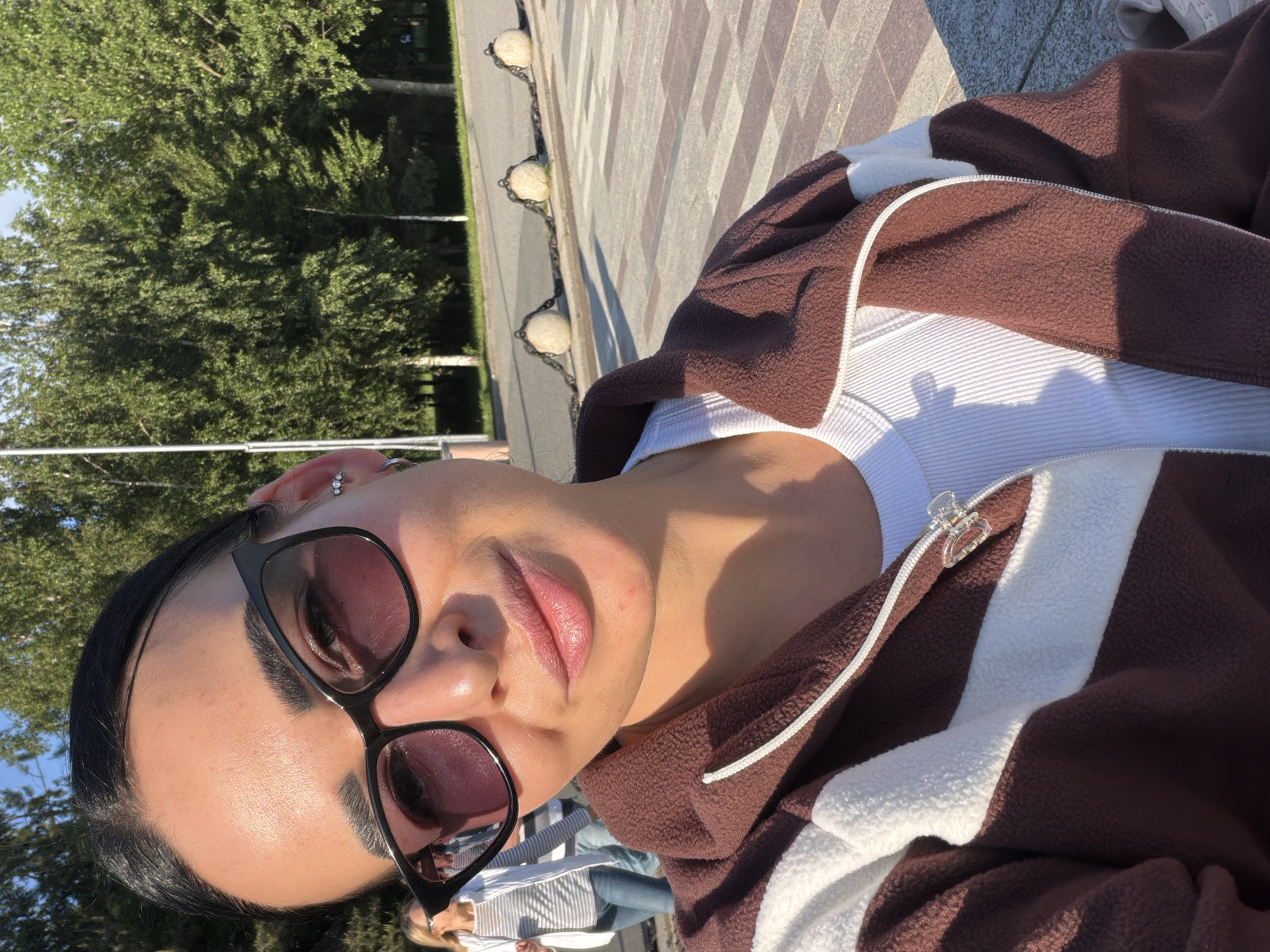 An outdoor, eye-level selfie captures a person looking directly at the camera, tilted to the right. The subject, with dark hair pulled back, wears large dark sunglasses, multiple earrings in their left ear, a white ribbed top, and a brown fleece jacket with white stripes and a zipper. A small clear hair clip is attached to the jacket's zipper pull.
The scene is brightly lit by natural sunlight, casting distinct shadows and highlighting the person's skin. In the background, lush green trees are visible, indicating a park or natural setting. A patterned paved walkway or promenade is also prominent, featuring a decorative chain barrier with spherical posts running along its edge. Another person, partially visible in the lower background, wears a white striped shirt and blue jeans, suggesting a public space. The clear skies and strong sunlight indicate good weather during daytime, possibly late morning or afternoon. No text is visible in the image. The location context is Saint Petersburg, Russia, aligning with an urban park or public walkway scene.