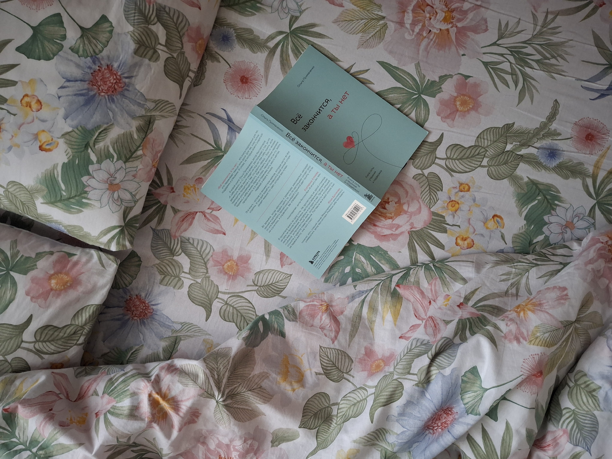 A top-down shot shows an open book resting on a floral-patterned bed with matching pillows. The book, titled "Всё закончится, а ты нет" (Everything will end, but you won't), is by Ольга Примаченко. The book cover is turquoise with a minimalist heart design. The book is open to show the text inside. The floral pattern on the bed consists of pink, white, and blue flowers amidst green foliage. The scene depicts a cozy, domestic setting, possibly in Chaikovsky, Russia, based on the book's language of origin.