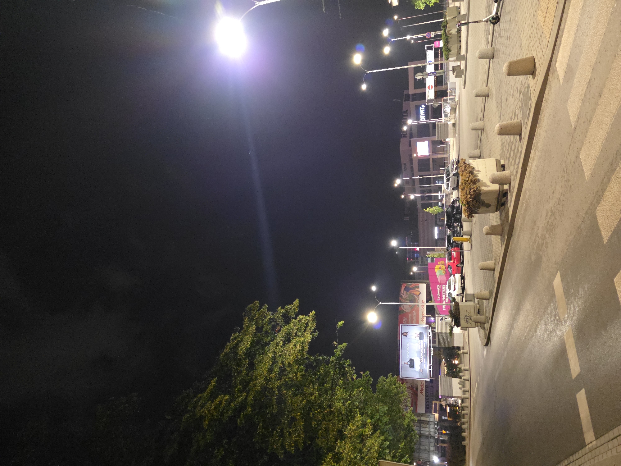 Captured at night in Warsaw, Poland, the image presents a street scene dominated by artificial lighting. The perspective is angled to the right, emphasizing the road and adjacent sidewalk.
On the right side of the frame, the sidewalk is paved with concrete blocks and features concrete bollards delineating the edge. A crosswalk with painted white stripes is visible on the pavement.
To the left of the road, trees with dense foliage partially obscure a commercial area. A large billboard advertising "Gino Rossi" and "Media Markt" is illuminated by streetlights. Other buildings are visible in the background, displaying commercial signage, including "MODIVO," "ACTION," and "OTYFIT".
Multiple streetlights illuminate the scene, casting a bright glow on the immediate environment and extending into the dark sky above. The overall impression is of a typical urban environment at night.