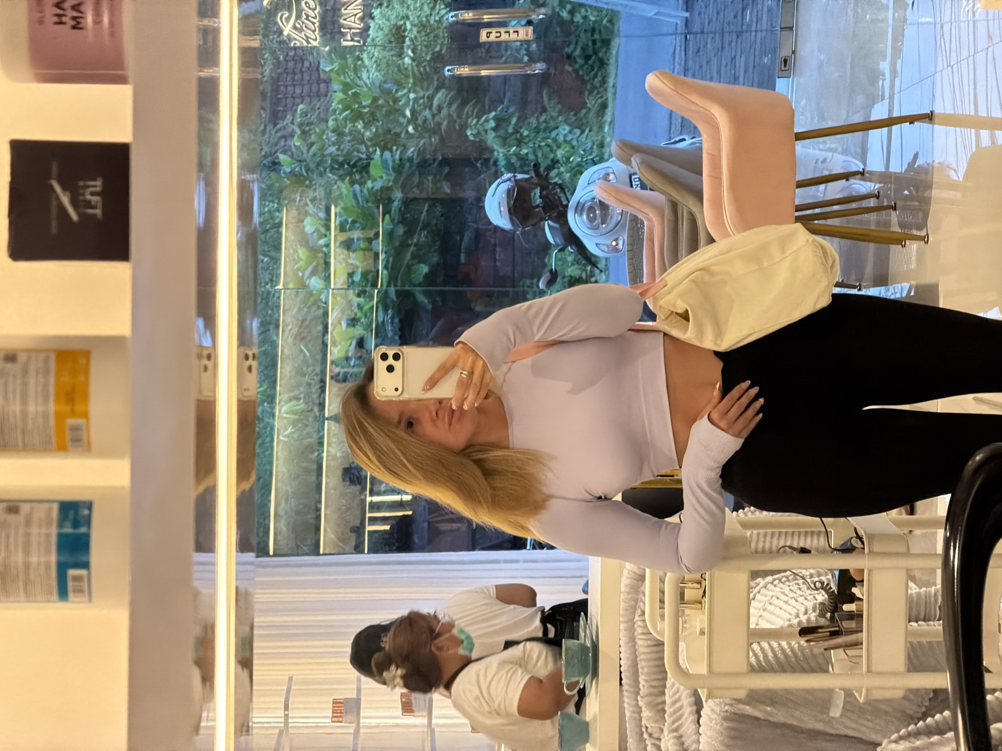 The image captures a selfie of a woman with blonde hair, wearing a white long-sleeved crop top and black leggings. She is holding a smartphone in front of her, presumably taking the picture.
In the background, visible through a glass partition, is a view of lush greenery and a scooter parked outside. Inside, another person wearing a face mask and a white shirt can be seen reflected. The woman is standing in a well-lit space that looks like a shop or studio. The presence of beauty products on the shelves suggests it may be a salon or beauty store. Also, a white fluffy towel, some beauty products and other equipment indicate the woman is in a beauty salon.
Based on the information provided, this image was taken in North Kuta, Indonesia.