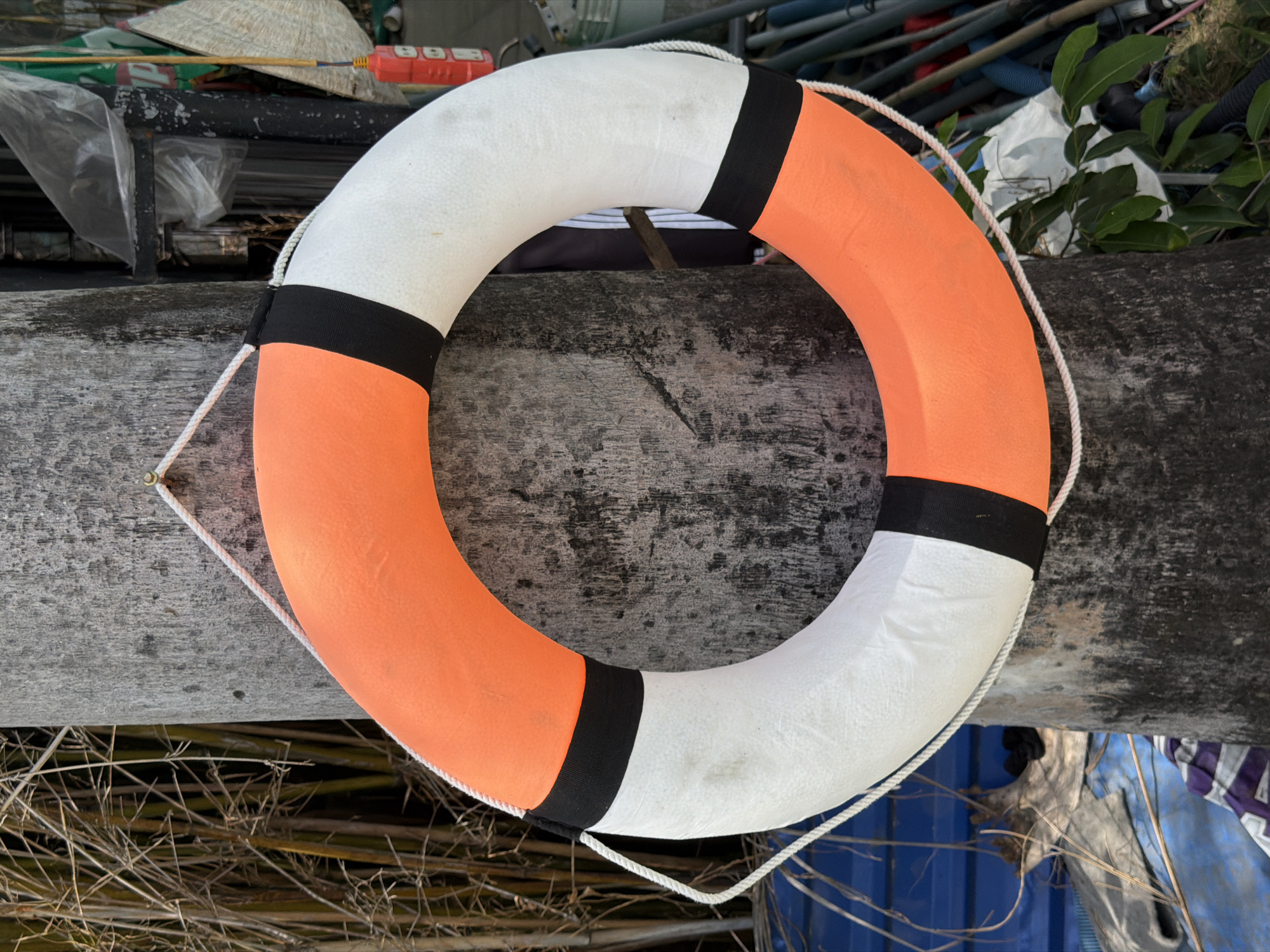 A life preserver with alternating orange and white sections and black bands rests on a weathered gray surface. A white rope is attached to the life preserver. The background includes vegetation, pipes, and other objects, suggesting an outdoor or industrial setting. The location is Nha Trang Ward, Vietnam.
