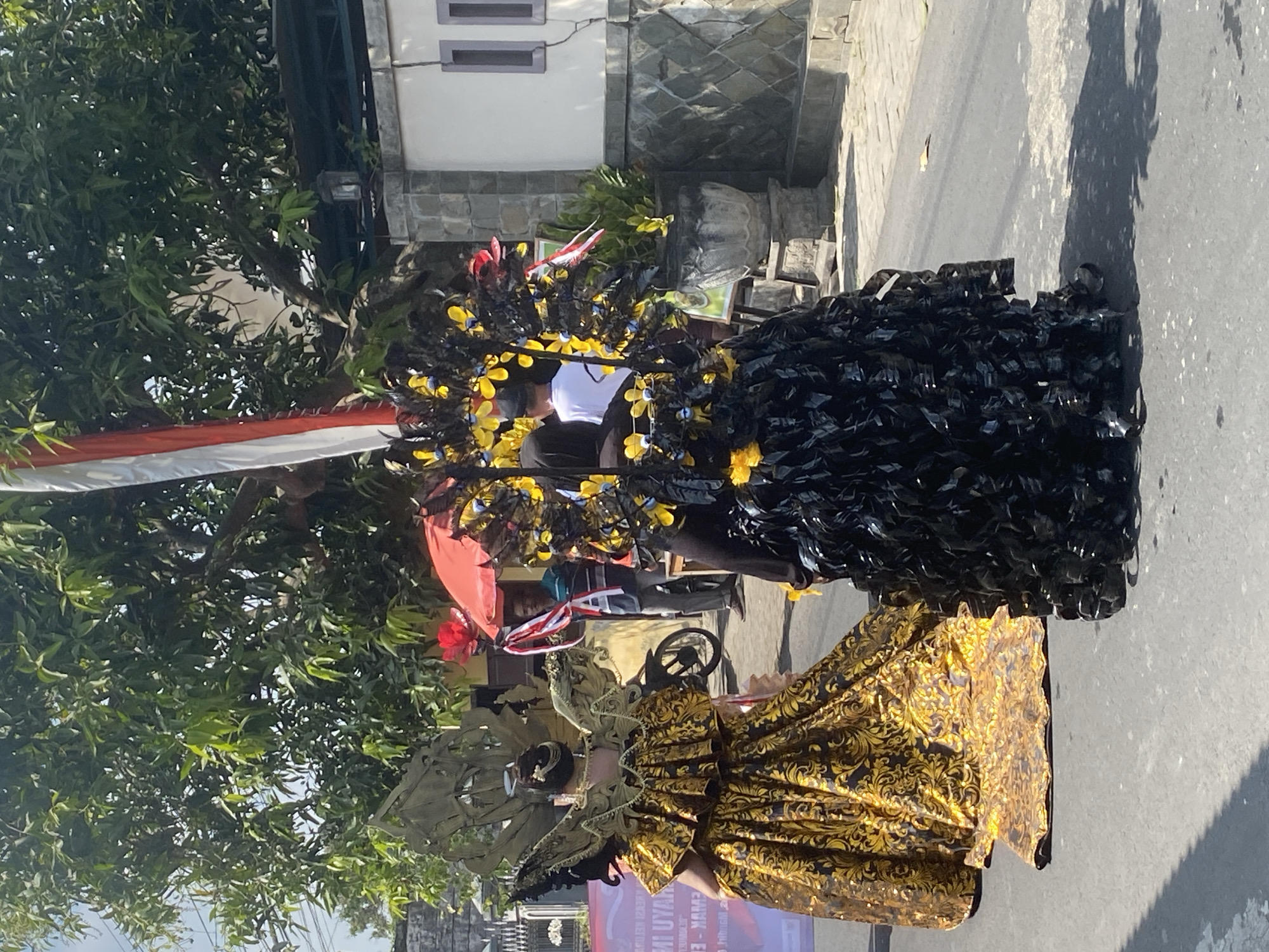 Two individuals in elaborate costumes are walking down a street in Kediri City, Indonesia. The person on the left wears a golden, ornate dress with a large, winged headpiece. The person on the right is dressed in a voluminous black costume adorned with yellow flowers. They are participating in what appears to be a street parade or festival. In the background, a motorcycle is parked, and people are watching from the side of the road. Trees and buildings line the street, suggesting an urban setting. The sun is shining, casting shadows on the asphalt. A red umbrella is visible, and a flag with red and white stripes is mounted on a pole.