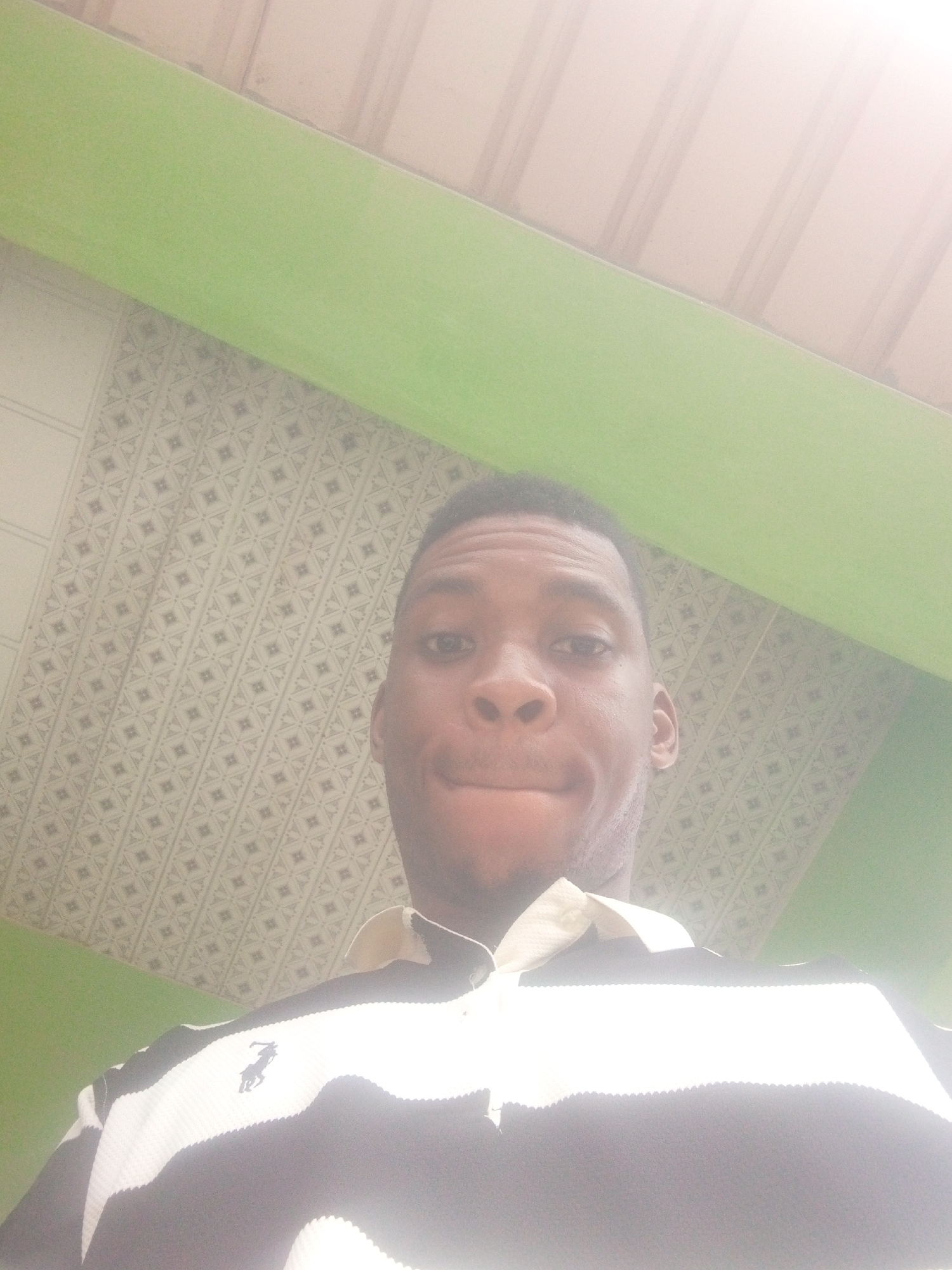 A medium, upward angle shot depicts a Black male in the city of Unknown, Unknown. He wears a striped polo shirt with a white collar and a dark emblem on the chest. Above him, there is a textured ceiling with a repetitive geometric pattern, bordered by a lime green beam. The overall lighting is bright and slightly hazy.