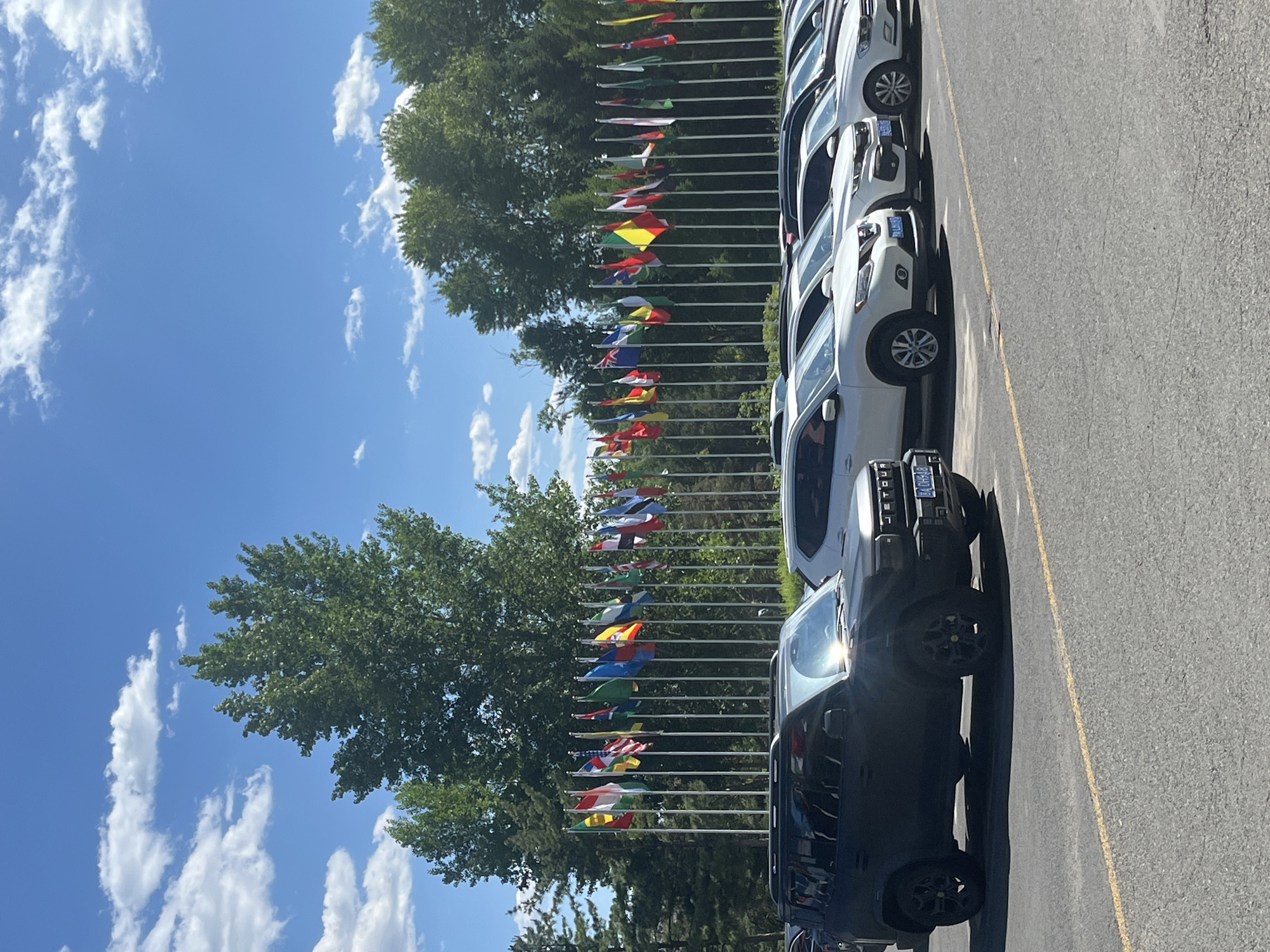 The image shows a row of cars parked along a yellow line on an asphalt surface. Behind the cars are a series of flagpoles, each bearing a different national flag. Trees are interspersed amongst the flagpoles. The sky above is blue with scattered white clouds. Based on the license plates, the location is most likely in Changchun City, China.
