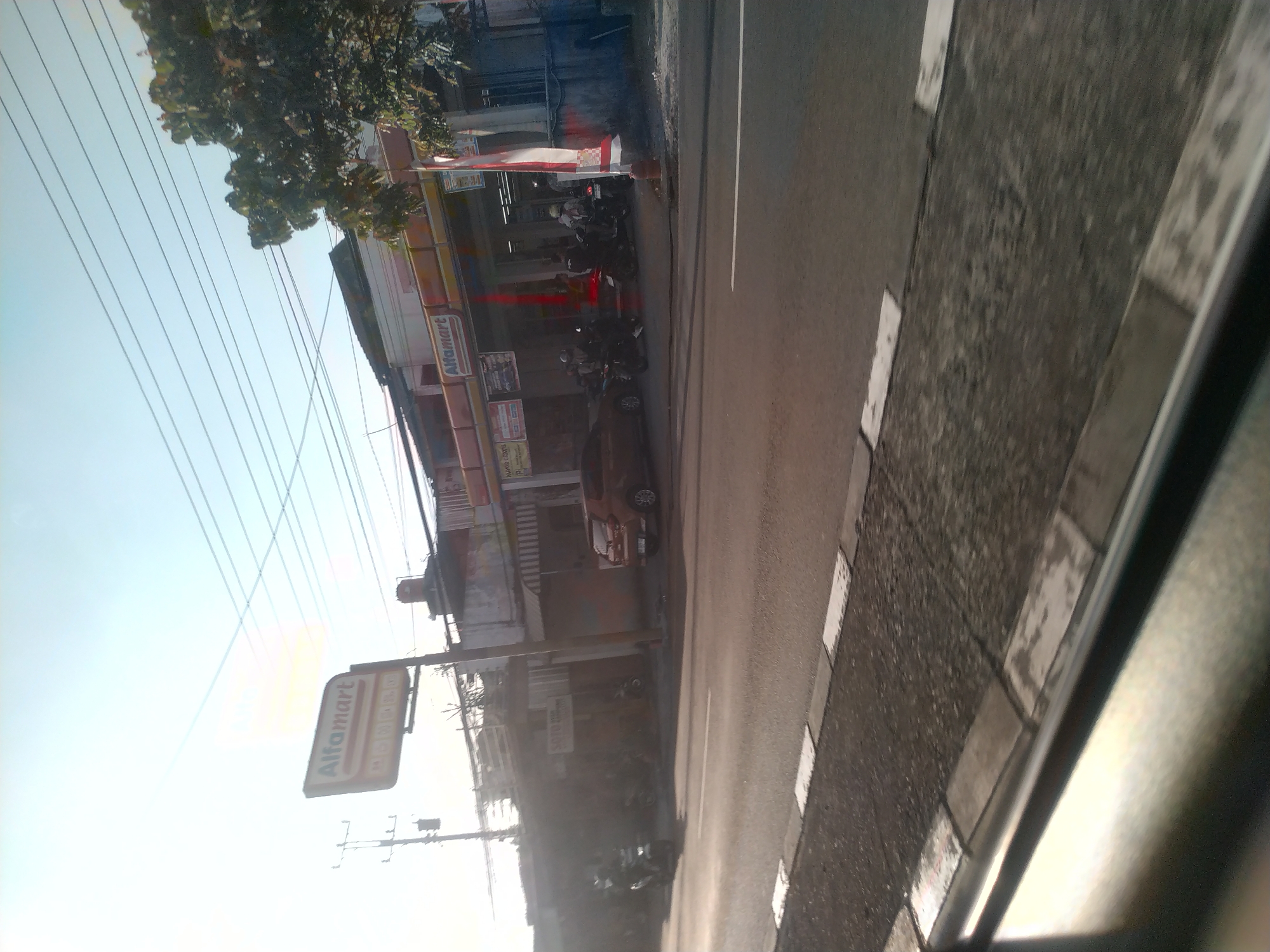 The image depicts a street scene in Teras, Indonesia, featuring an Alfamart convenience store. A red car is parked in front of the store, and several motorcycles are visible on the sidewalk. The sky is bright and clear with power lines strung across it. The foreground shows a roadside curb and a portion of a road. The overall atmosphere is that of a typical commercial street in Indonesia, with the Alfamart store being the central focus.