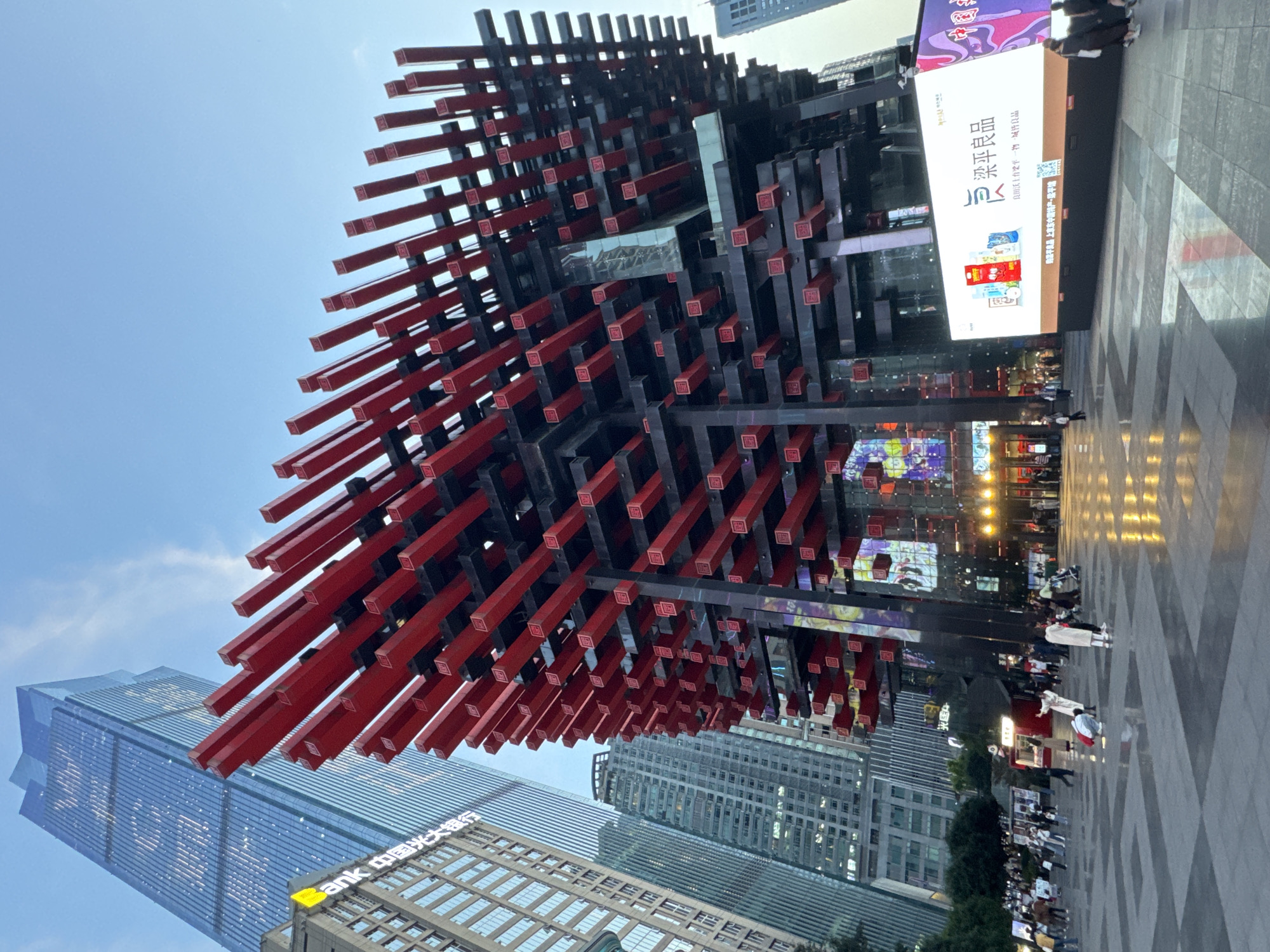 The image captures the Raffles City Chongqing, a complex in Yuzhong District, Chongqing, China, dominated by a massive, intricate art installation. The sculpture is comprised of numerous red and black beams arranged in a cube-like structure. It’s positioned in a plaza. People mill around in the plaza. The environment includes modern high-rise buildings, commercial displays, and city lights. One side of the sculpture is adjacent to an advertising display. The sky is a light blue.
