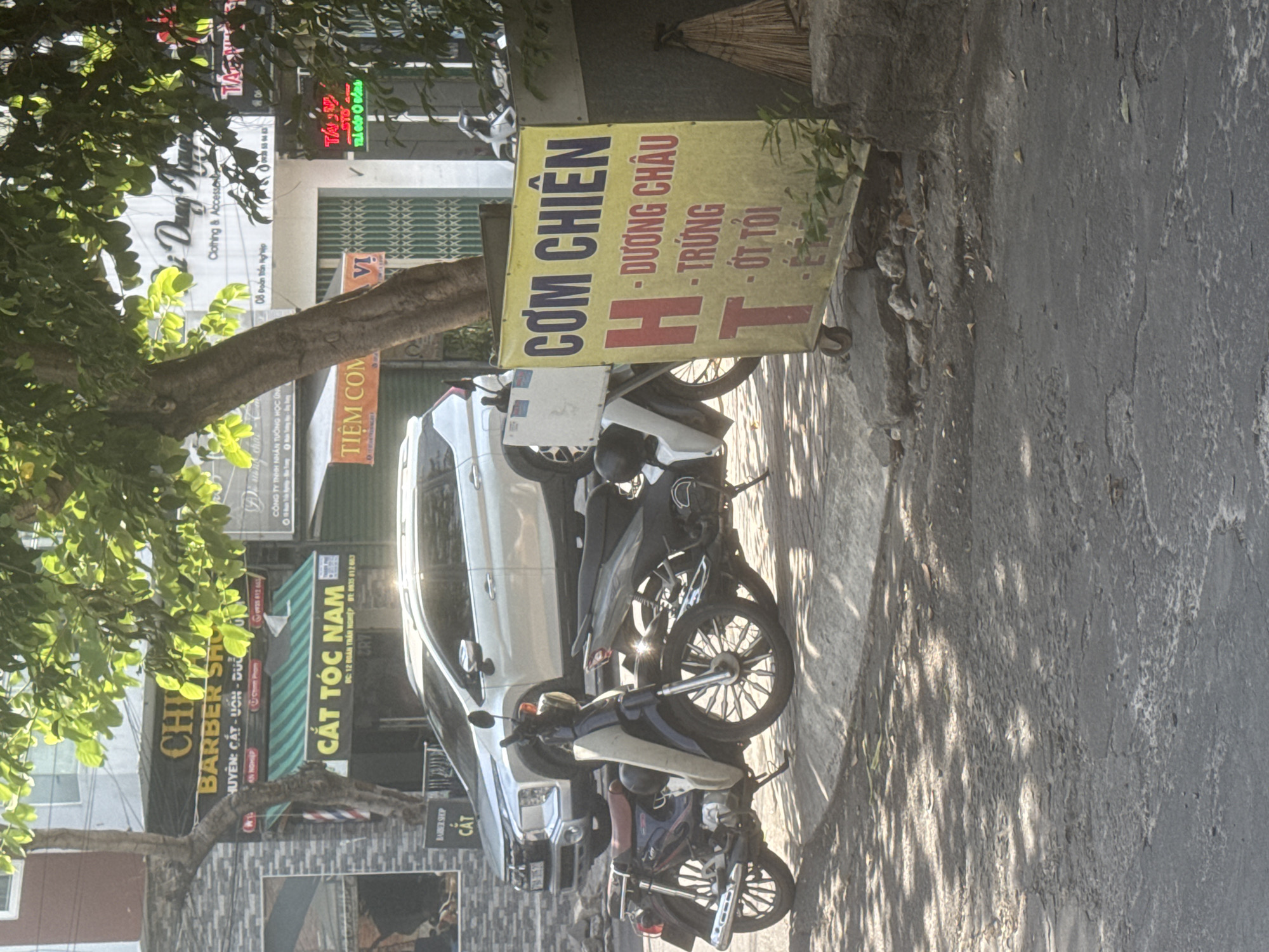 The image depicts a street scene in Bac Nha Trang Ward, Vietnam. A silver SUV is parked on the right side of the frame, partially obscuring two motorcycles parked in front of it. The first motorcycle, on the left, is dark blue with a brown seat and a light-colored front panel. The second motorcycle, on the right, is dark gray. A yellow sign with Vietnamese text is prominently displayed in the foreground, advertising "COM CHIEN" (fried rice) and listing ingredients such as "Duong Chau" (Yangzhou), "Trung" (egg), and "Ot Toi" (chili garlic). The sign also features large red letters "H" and "T". Behind the vehicles and signs, a building with a barber shop sign "CAT TOC NAM" (Men's Haircut) and other storefronts are visible. The area is shaded by trees with sunlight dappling the pavement. The overall environment appears to be an urban street with commercial establishments.