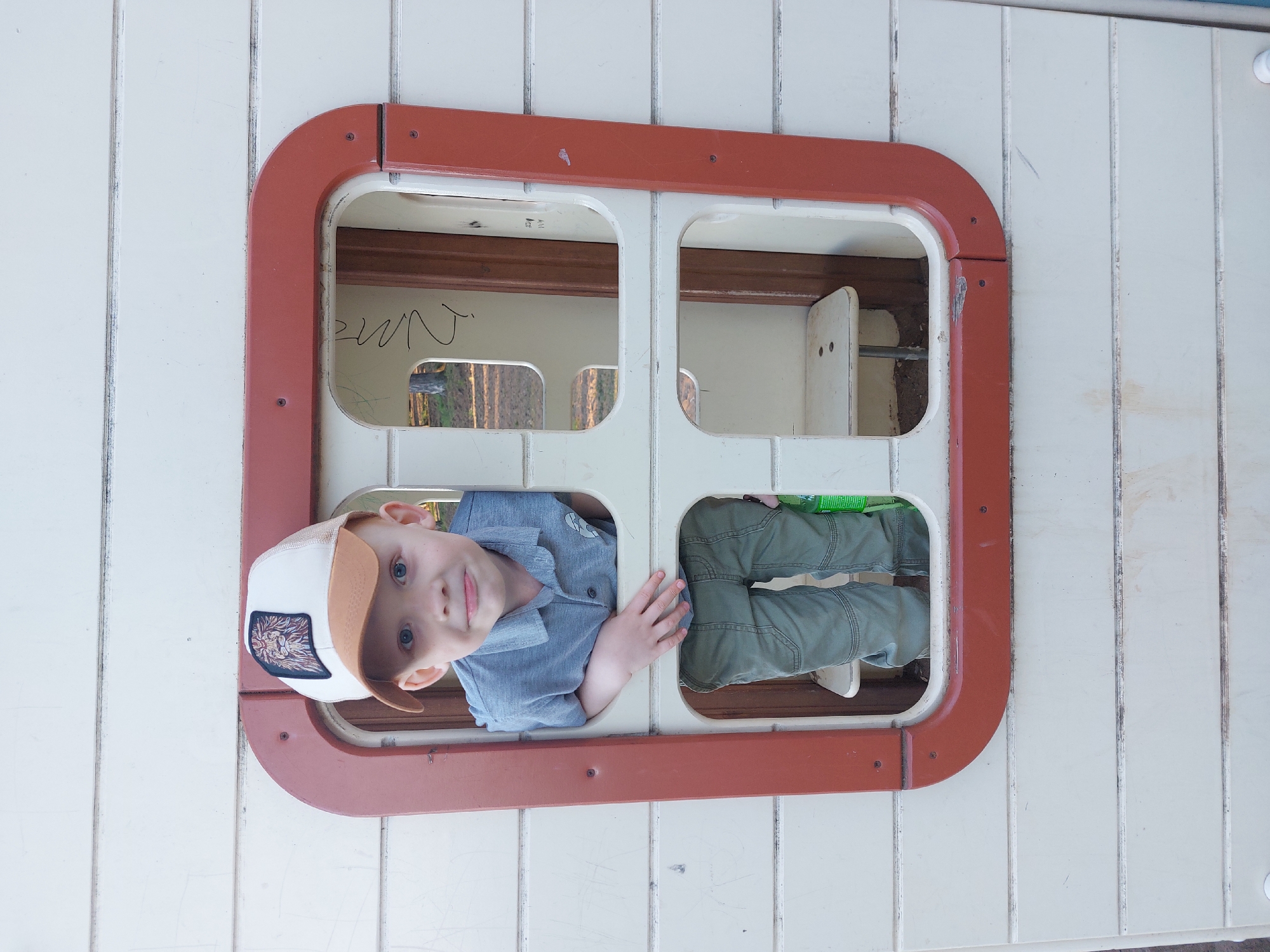 A young child is framed in a rectangular window with rounded corners, divided into four panes. The window is set in a wall of vertical boards, possibly part of a playhouse or structure. The child is wearing a light-colored baseball cap with a brown bill and a patch depicting a lion or similar animal. He has blue eyes and is wearing a light blue collared shirt. His face is visible through the upper left pane, and his legs, clad in green pants, are visible through the lower right pane.
The context of the city of Chaikovsky, Russia is not directly visible within the image.